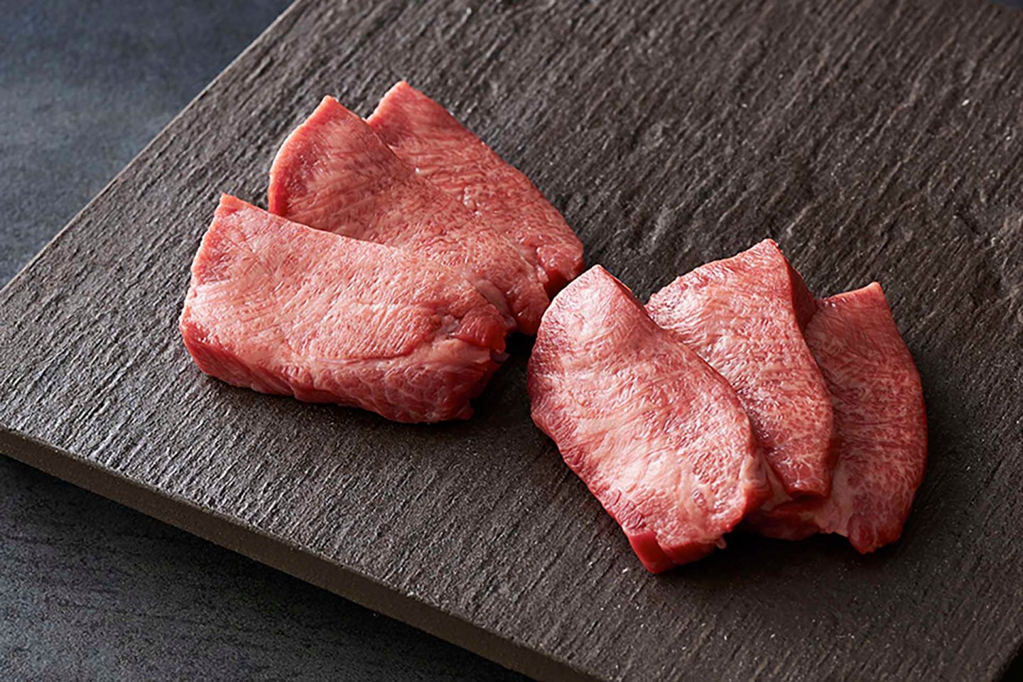 Four slices of raw beef tongue are arranged on a dark textured surface.