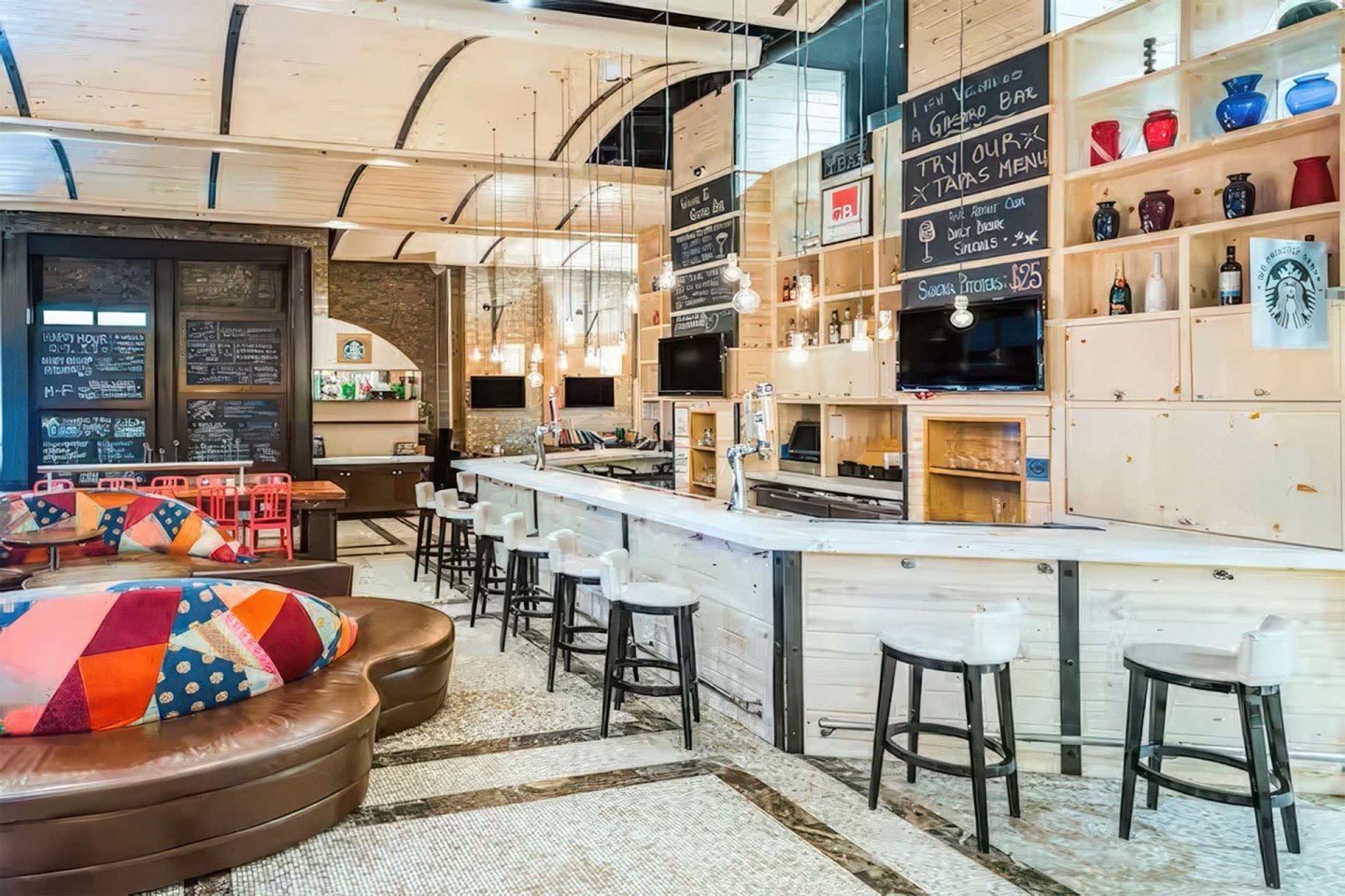 A modern bar with light wood paneling, high stools, chalkboard menus, colorful pillows on brown leather sofas, shelves with vases and bottles, and hanging pendant lights in a bright, inviting space.