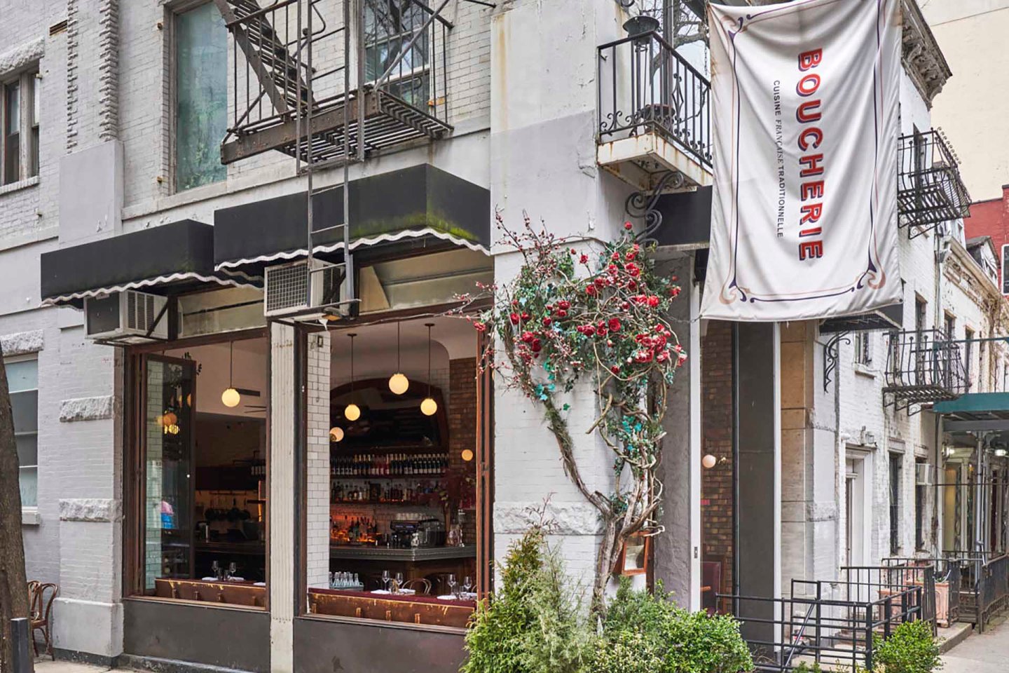 Street view of a restaurant exterior with a sign reading "Boucherie." The entrance features large windows, black awnings, and a vine with red flowers climbing the building. Lights are visible inside, along with bottles lined on shelves.