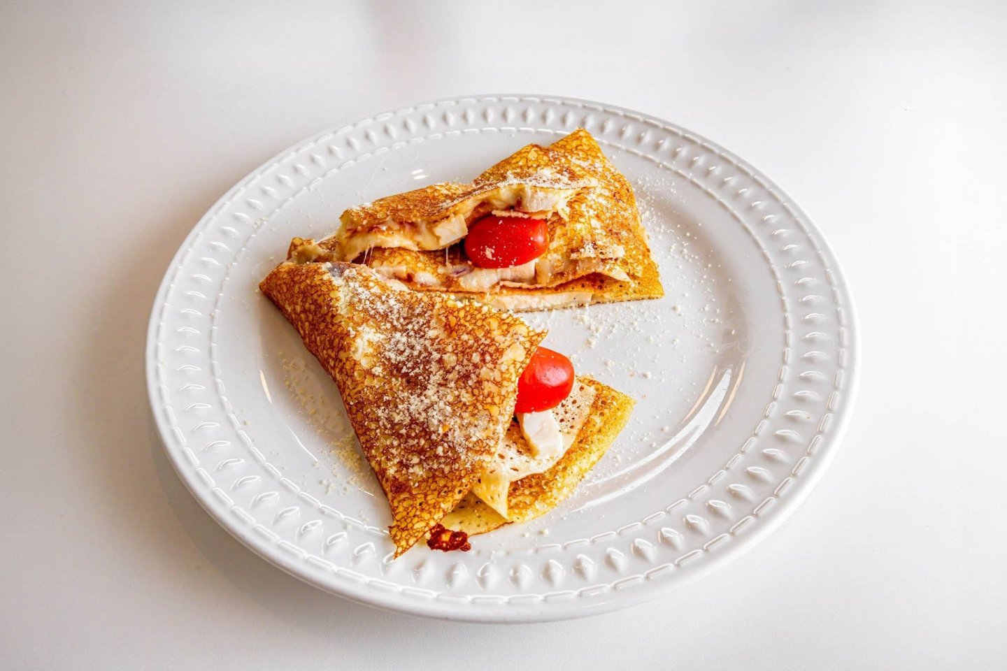 Two folded crepes filled with cherry tomatoes and creamy cheese, sprinkled with grated parmesan, are served on a white decorative plate against a plain white background.
