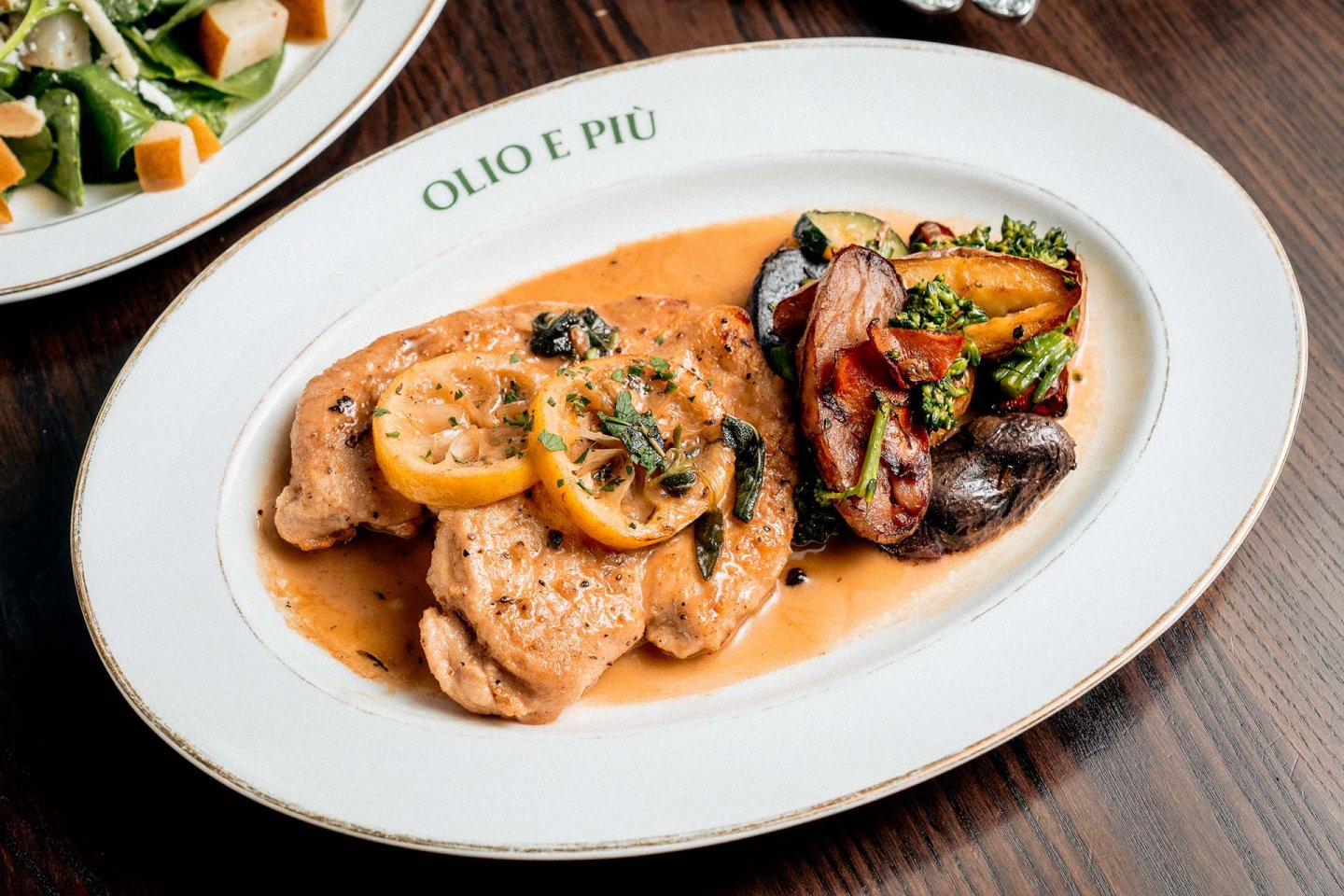 Grilled chicken breasts with lemon slices and sauce, served with roasted potatoes, broccoli, and carrots on a white plate labeled “OLIO E PIÙ” on a wooden table. A side salad is partially visible.