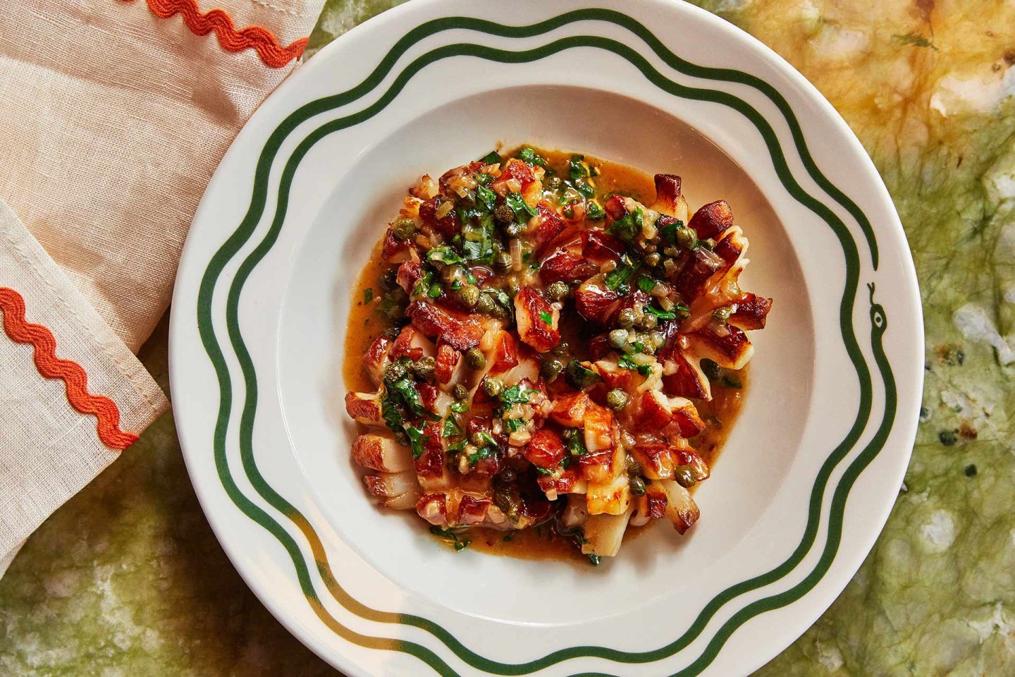 A plate with a neatly arranged dish of chopped octopus in a rich tomato-based sauce, garnished with fresh herbs, sits on a patterned green and white plate atop a marbled surface with a folded napkin nearby.