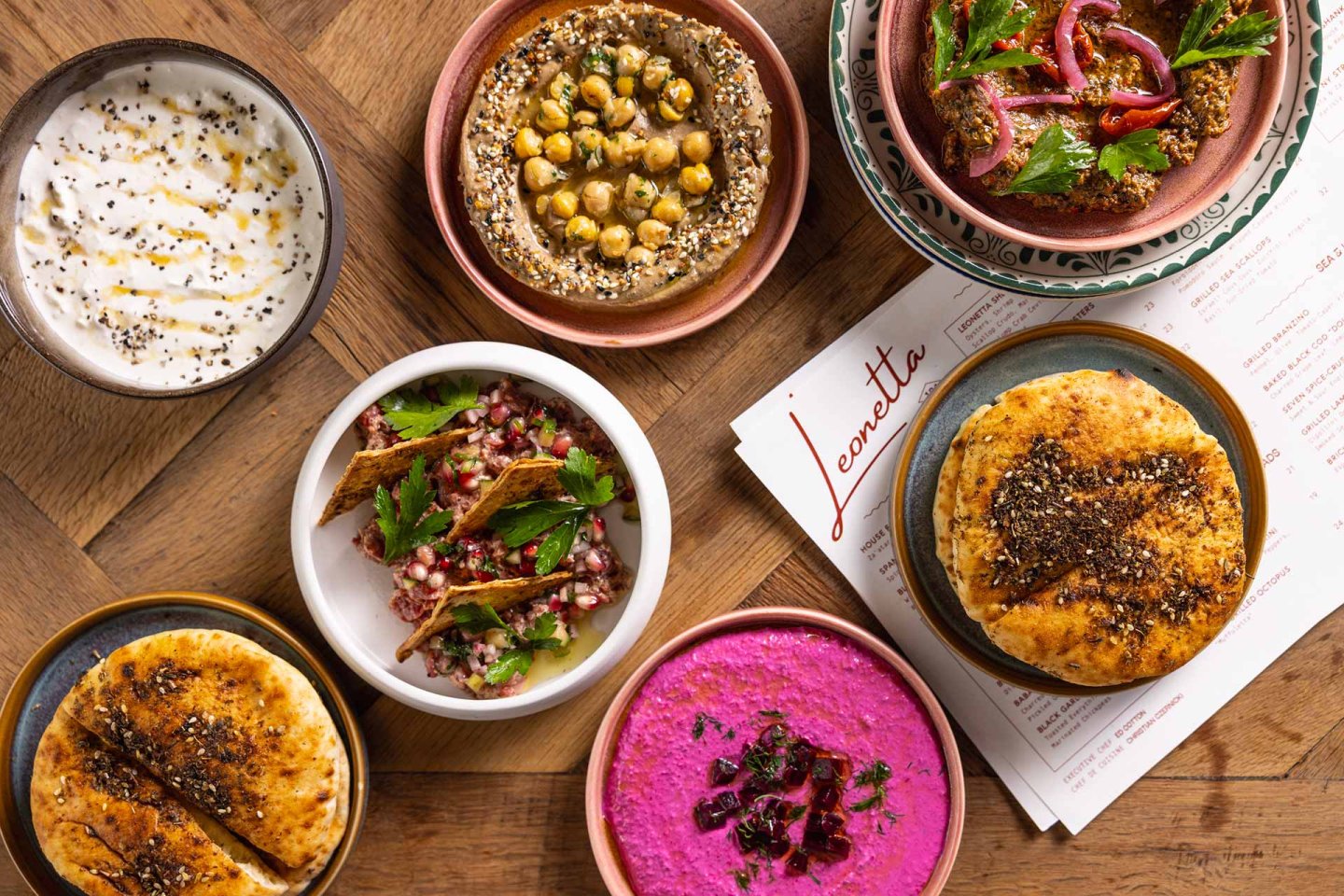 Assorted Middle Eastern dishes including pita, hummus, labneh, beet dip, tabbouleh, and baba ganoush in colorful bowls on a wooden table, with part of a menu visible.