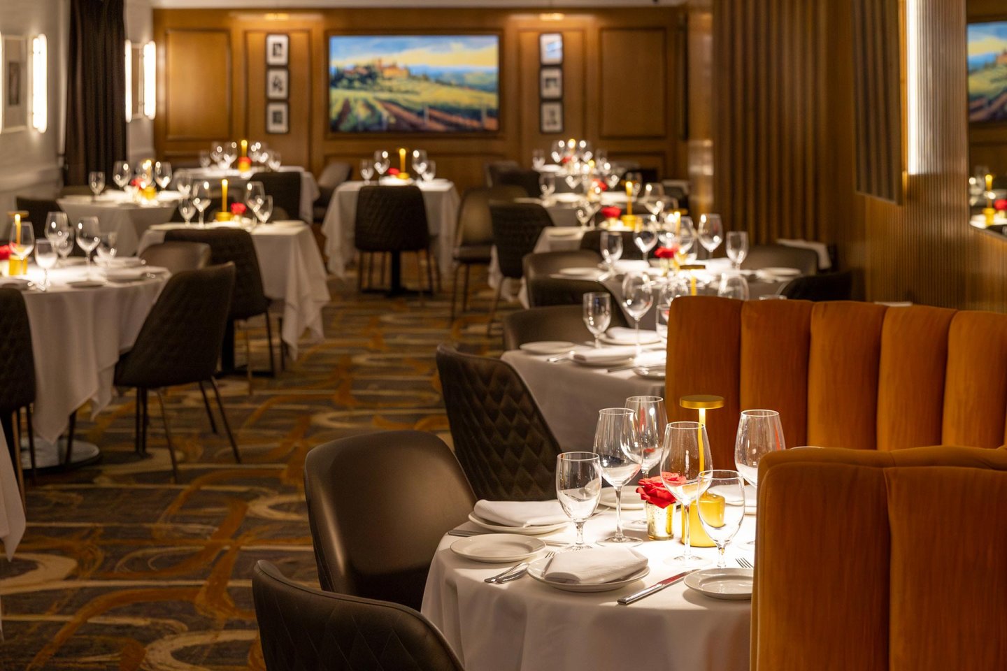 Elegant restaurant interior with white tablecloths, set tables with wine glasses, candles, and red flower centerpieces. Warm lighting and paintings on wood-paneled walls create a cozy, upscale ambiance.