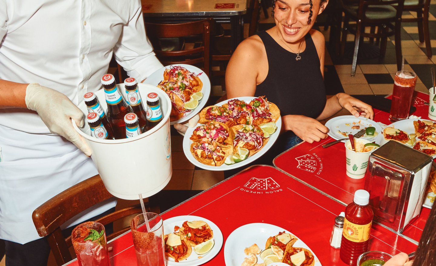 A server carries a bucket of bottled drinks and plates of tacos to a table of guests in a lively taqueria. Bright red tables are covered with tacos, cocktails, salsa, and lime wedges as diners gather for a festive meal in a warm, bustling restaurant.