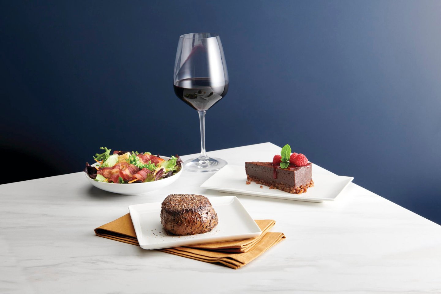 A sophisticated meal arrangement featuring a glass of red wine, a fresh salad in a bowl, a seasoned steak on a plate, and a slice of chocolate cake garnished with raspberries and mint on another plate, set on a white marble table.