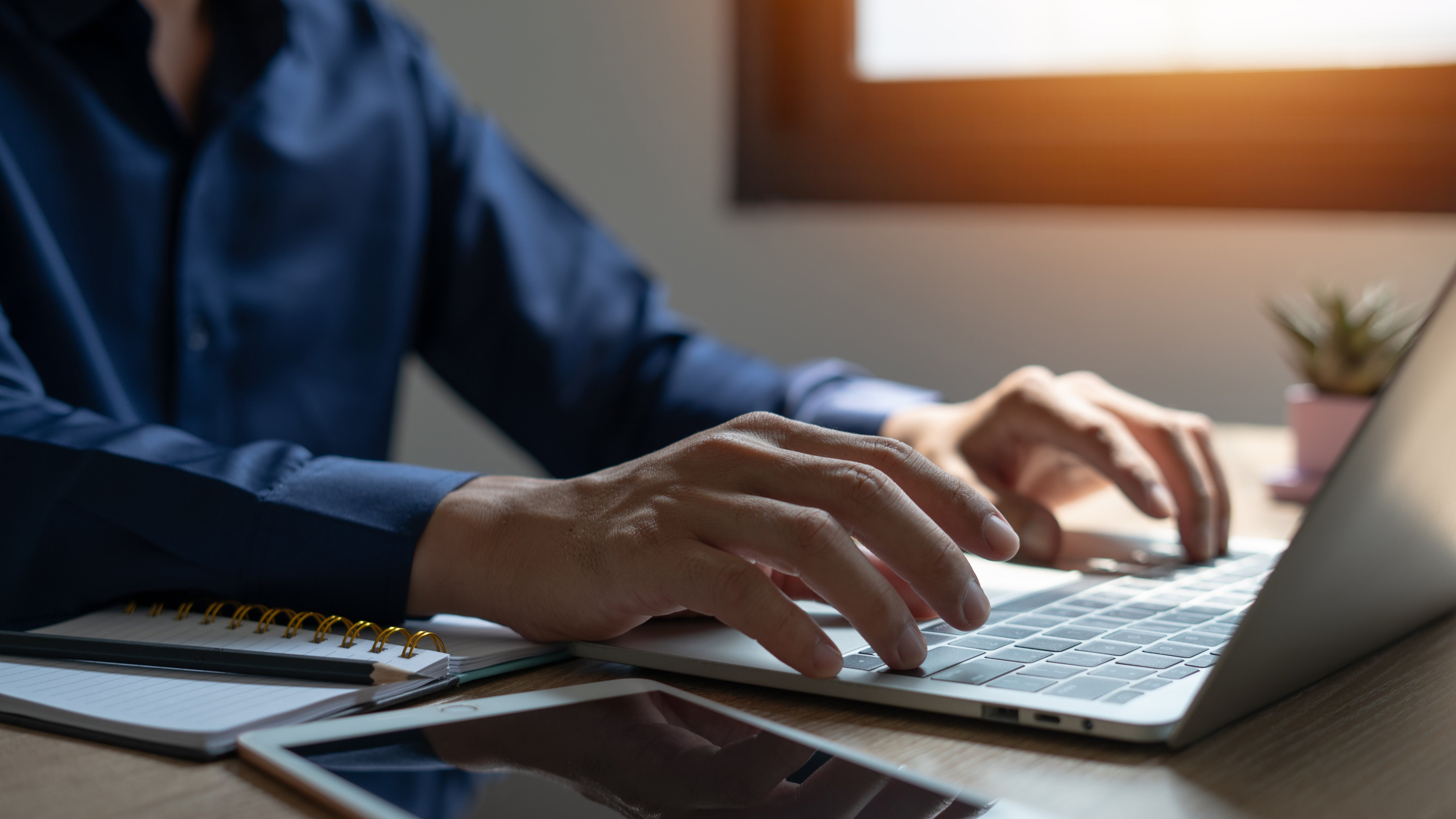 A man is typing on a laptop in front of a window. The laptop is open and the man's hands are on the keyboard. There are several notebooks and a tablet on the desk