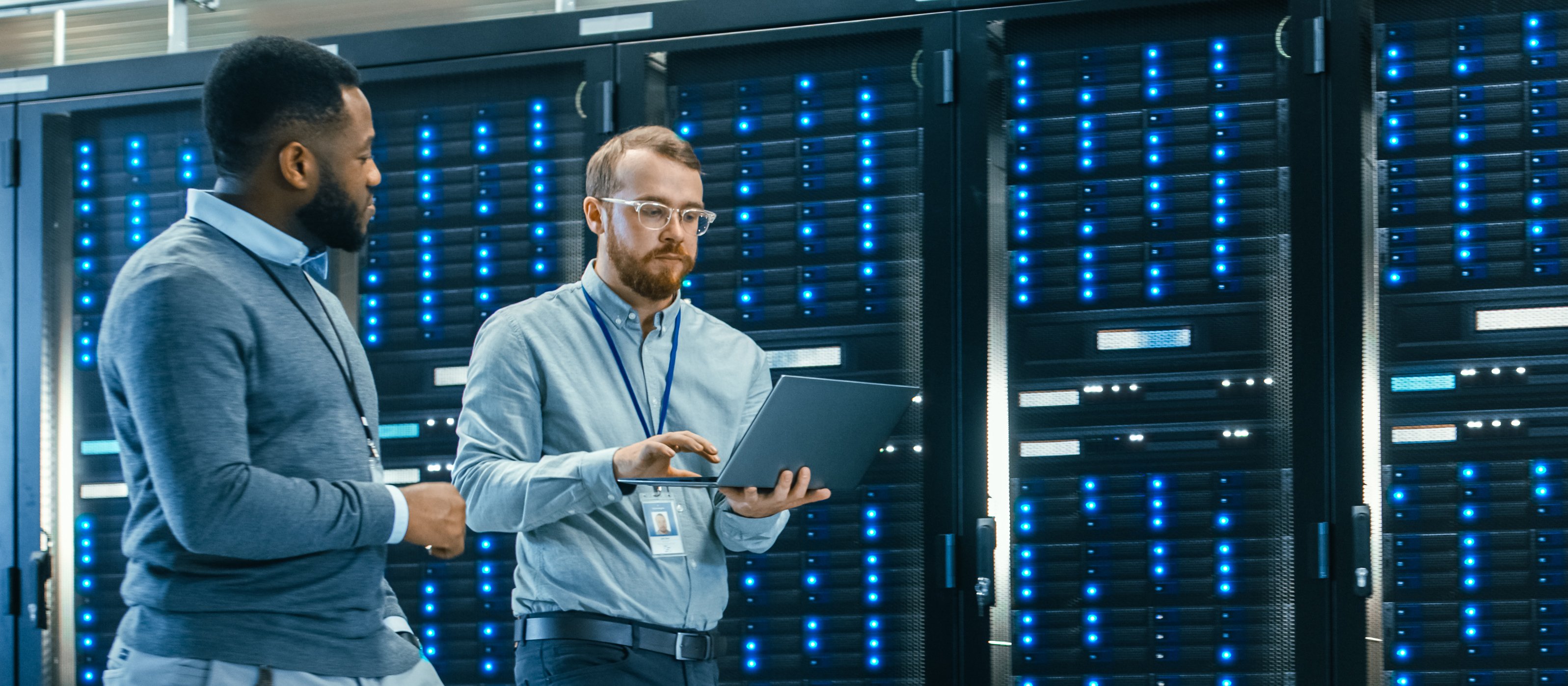 IT Technician with a Laptop Computer and Black Male Engineer Colleague are Talking in Data Center while Walking Next to Server Racks. Running Diagnostics or Doing Maintenance Work.