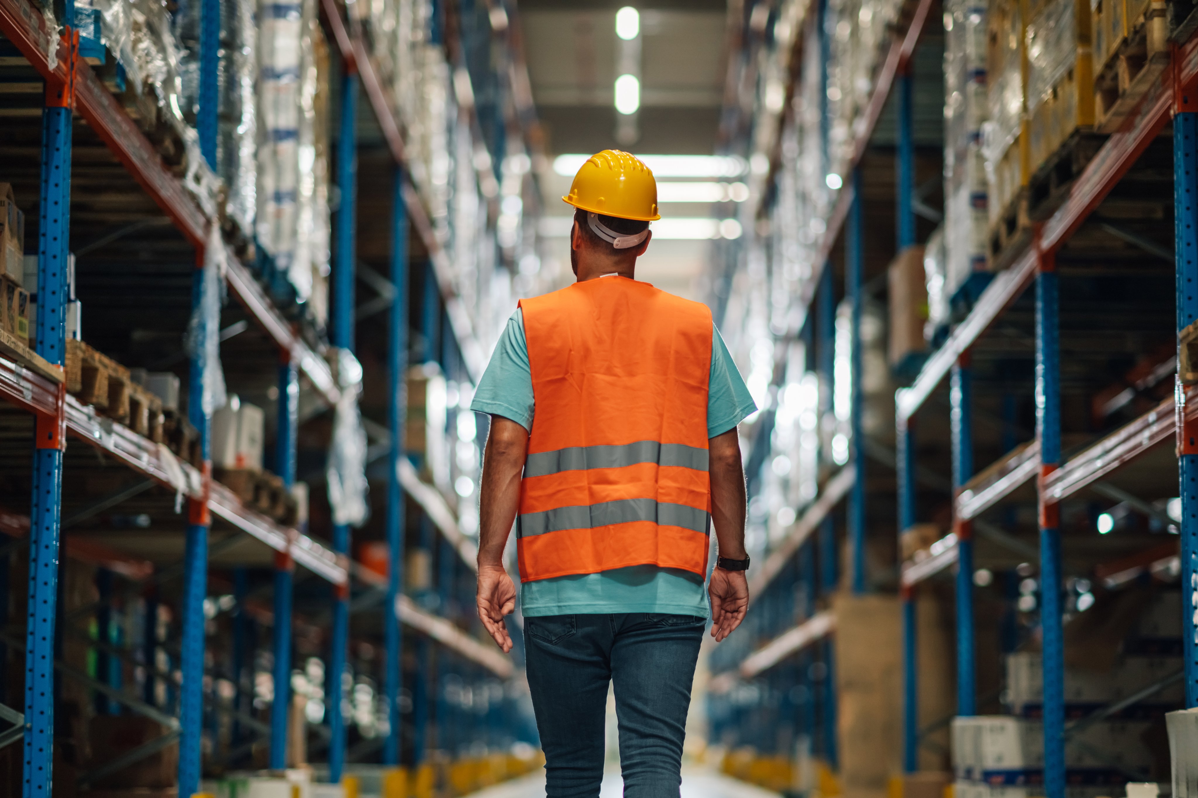 Warehouse worker wearing safety vest and helmet walking away through large distribution warehouse between shelves full of goods, checking inventory and managing logistics