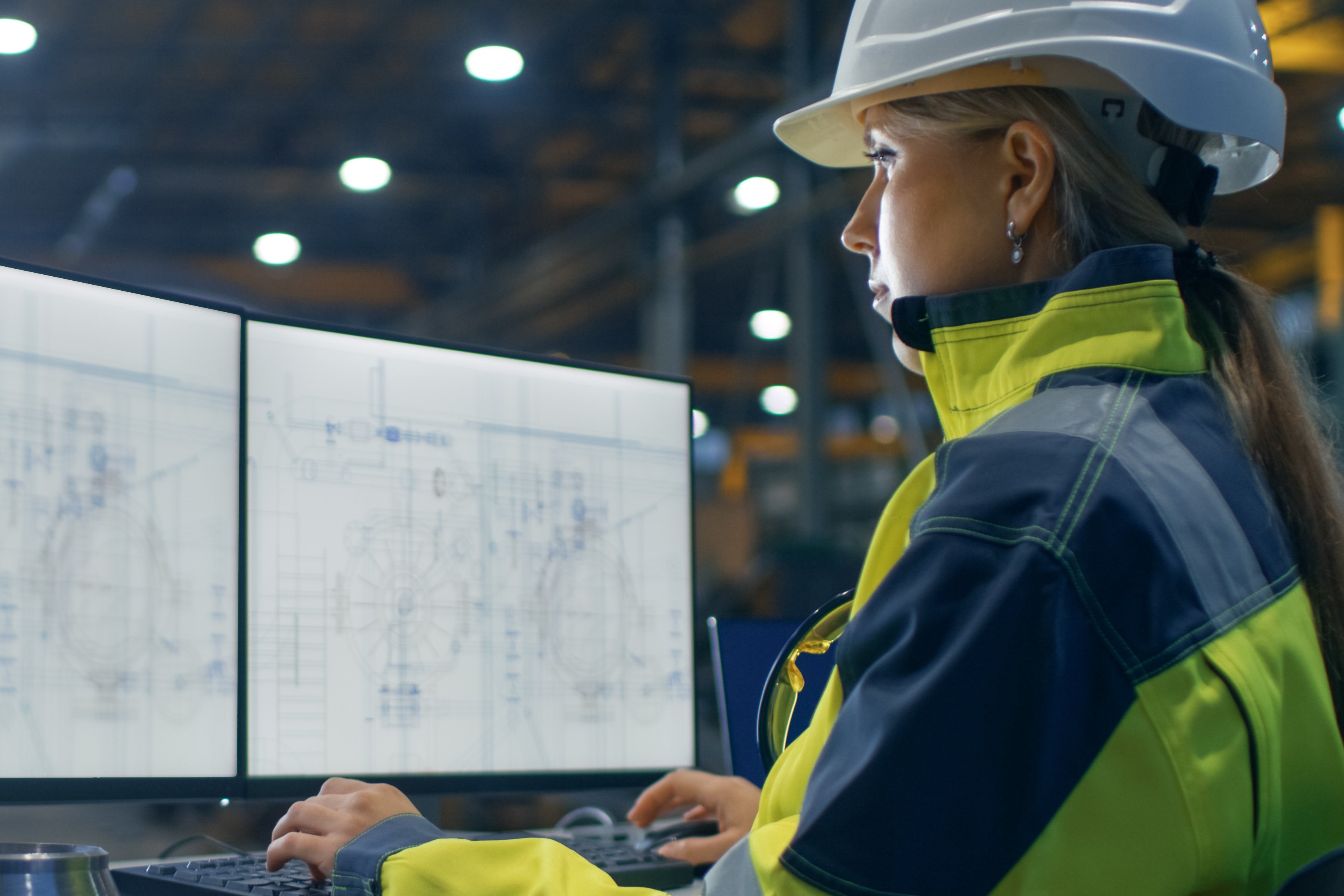 Inside the Heavy Industry Factory Female Industrial Engineer Works on the Personal Computer with Green Mock-up Screen. Secondary Monitor Shows Blueprints.