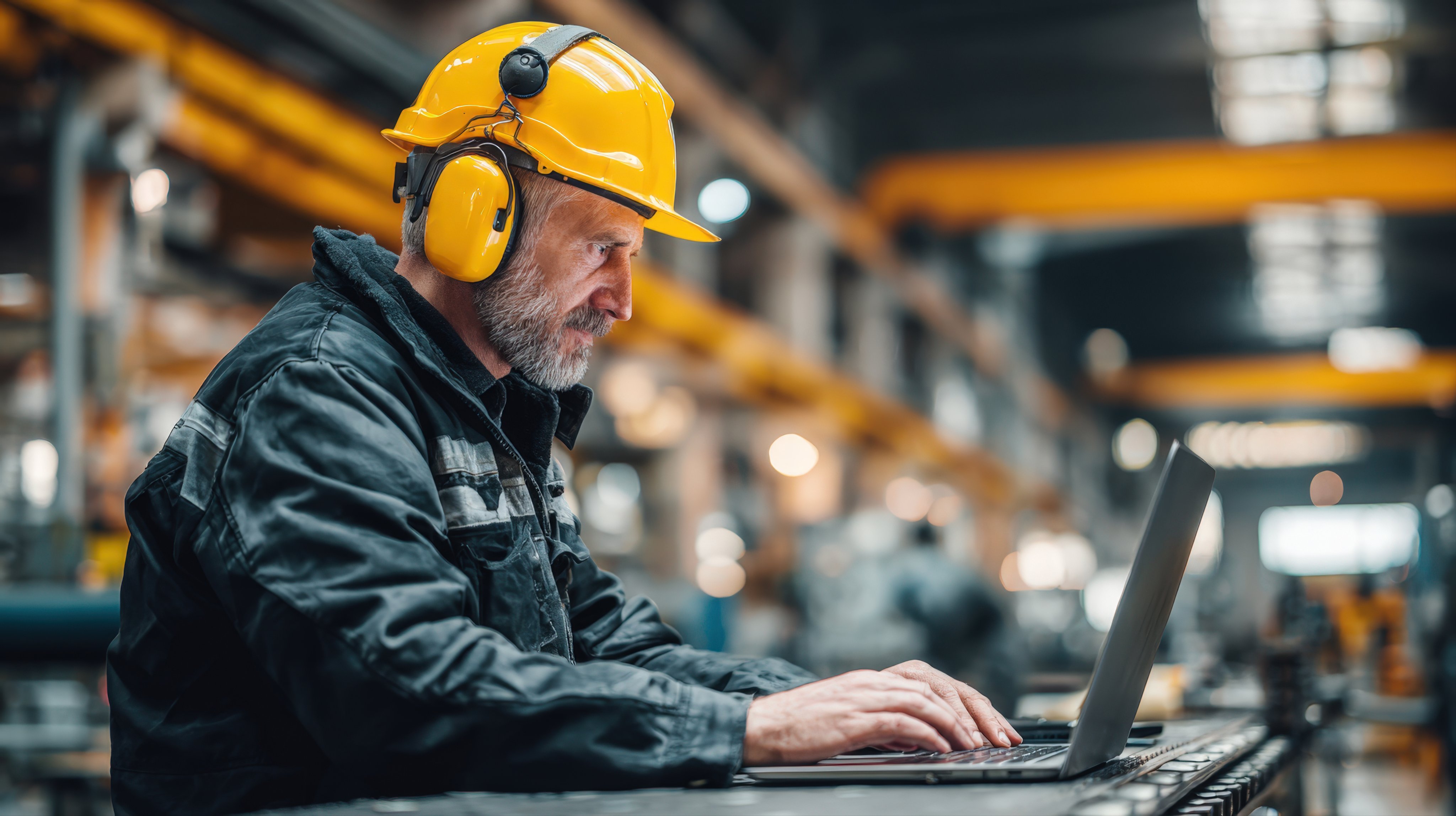 A focused, mature engineer in a hard hat and hearing protection works on a laptop on the factory floor. He is a professional worker, combining technology with hands-on industrial work.