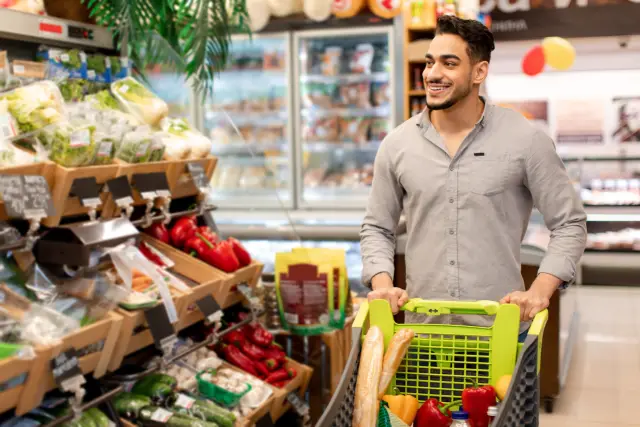 Happy Arabic Buyer Guy Shopping Groceries Walking With Cart Full Of Food In Modern Supermarket Indoor. Customer Purchasing Products On Weekend. Commerce And Consumerism
