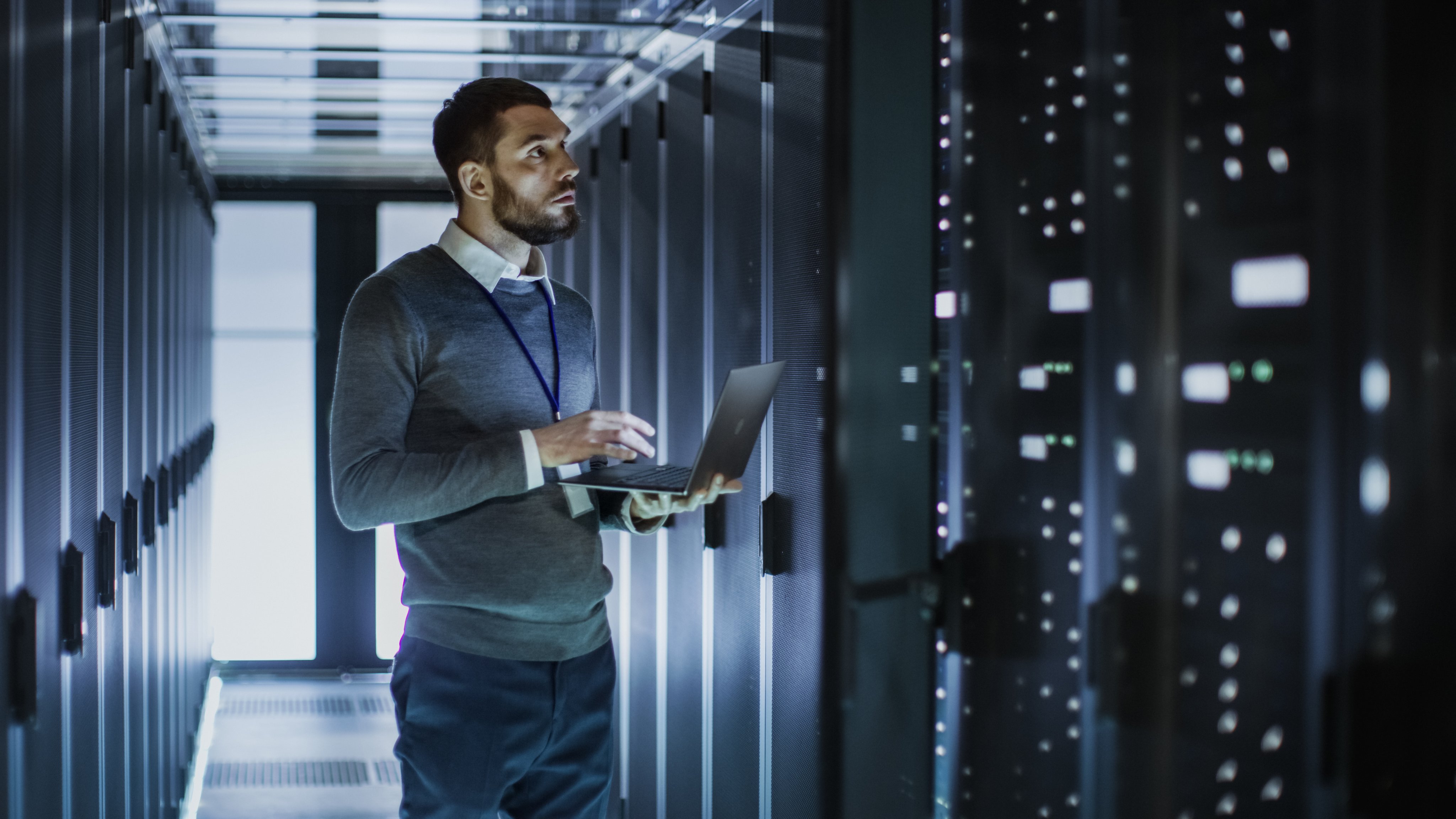 IT Technician Works on Laptop next to a Server Cabinet in Big Data Center. He Runs Diagnostics and Maintenance, Sets System Up.