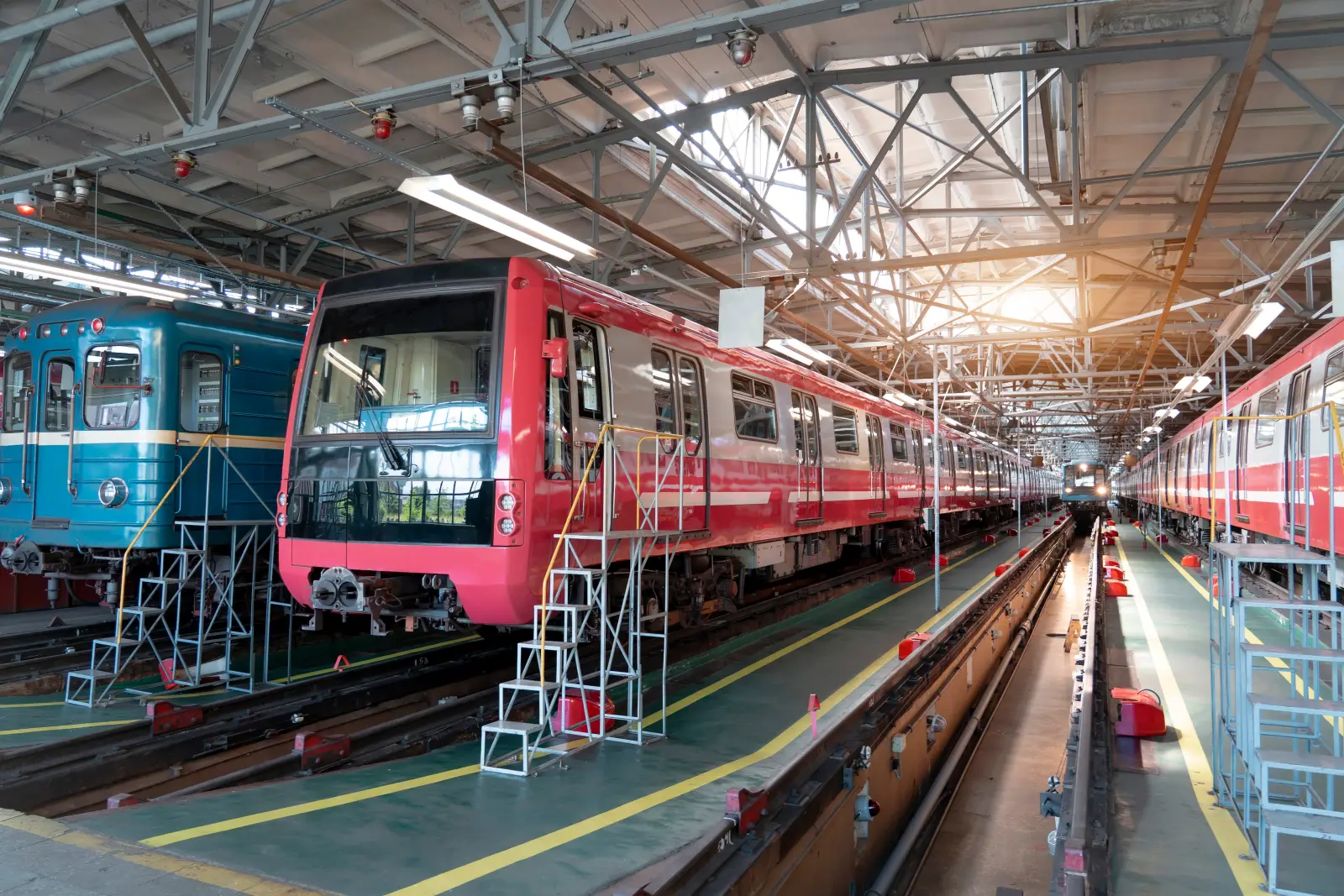 Several subway trains are parked on tracks within a spacious industrial maintenance facility. A modern red and white train is prominent, with an older blue train nearby