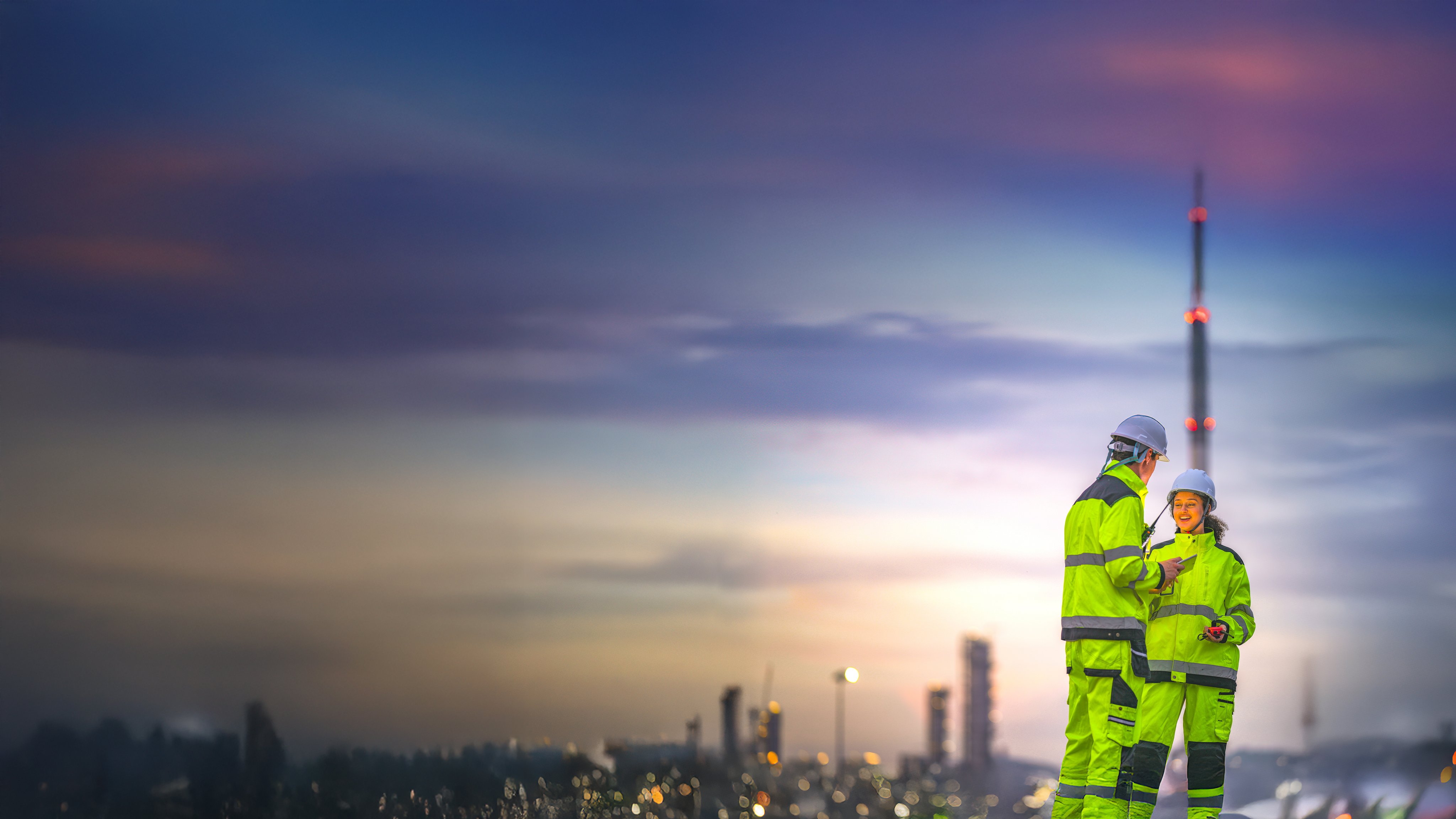 Two engineers in high-visibility suits and helmets review data on a tablet while standing in front of a refinery at sunrise, indicating teamwork and safety in a heavy industry zone.