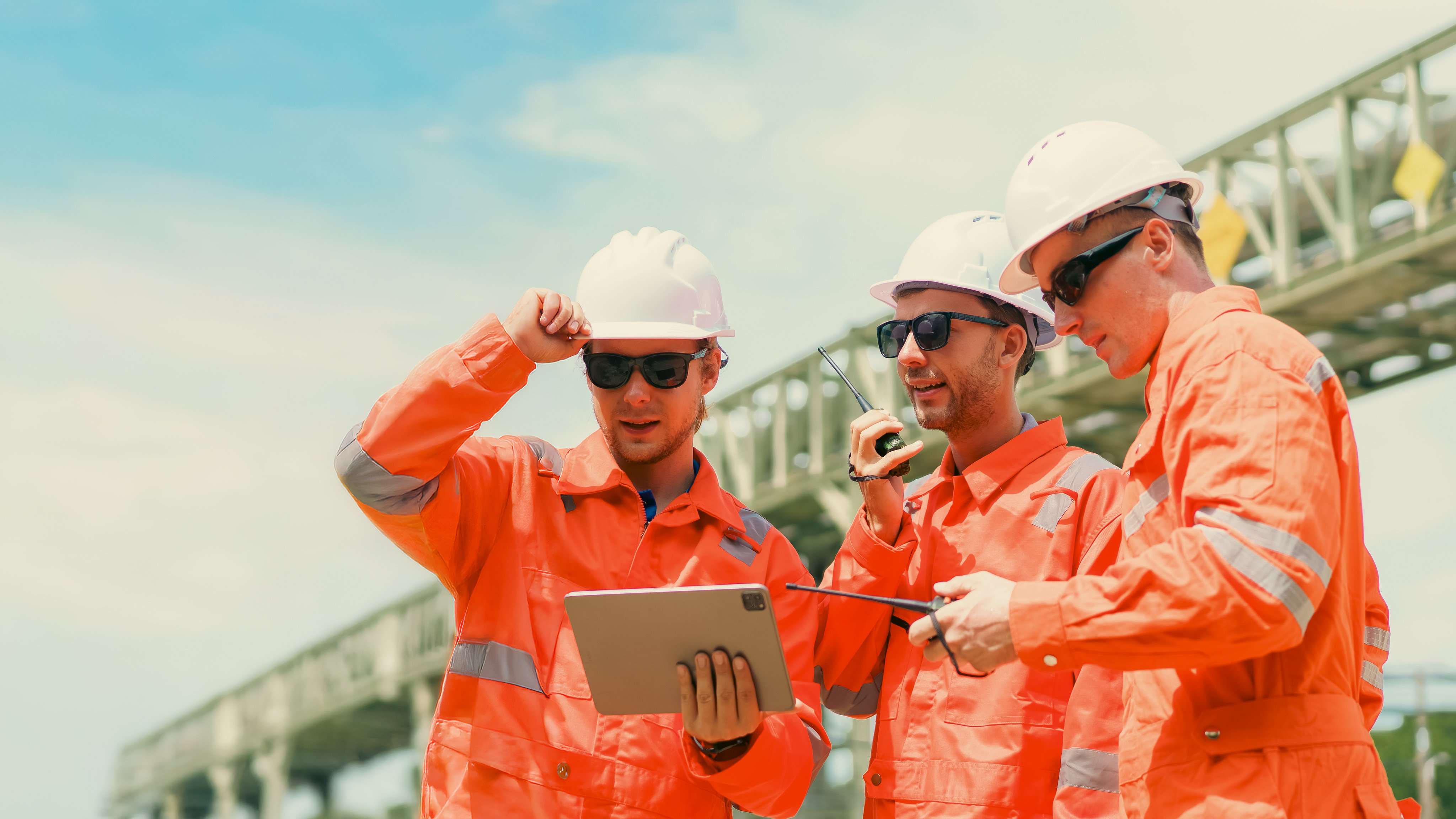 Team of engineers wearing a safety vests and helmets inspect refinery pipelines while discussing and reviewing an operation plan on a tablet.