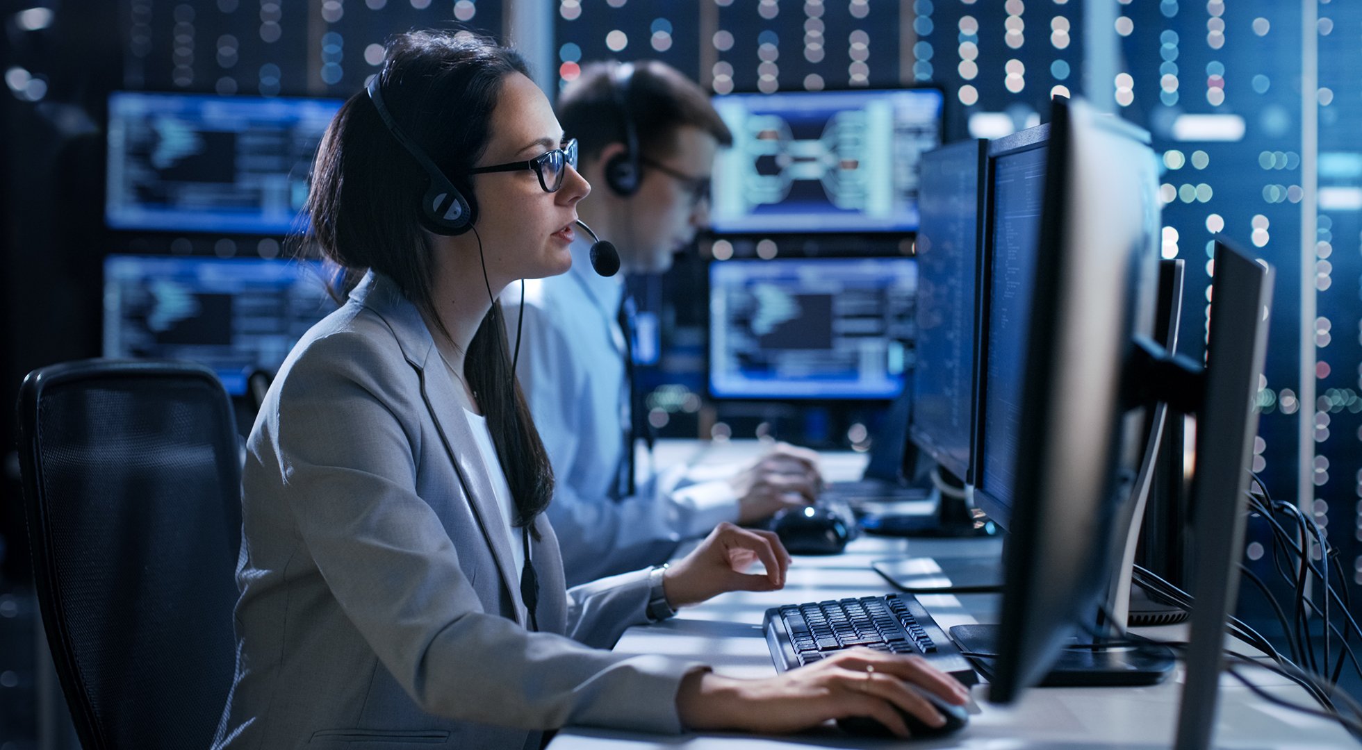 Female working in a Technical Support Team Gives Instructions with the Help of the Headsets. In the Background People Working and Monitors Show Various Information.