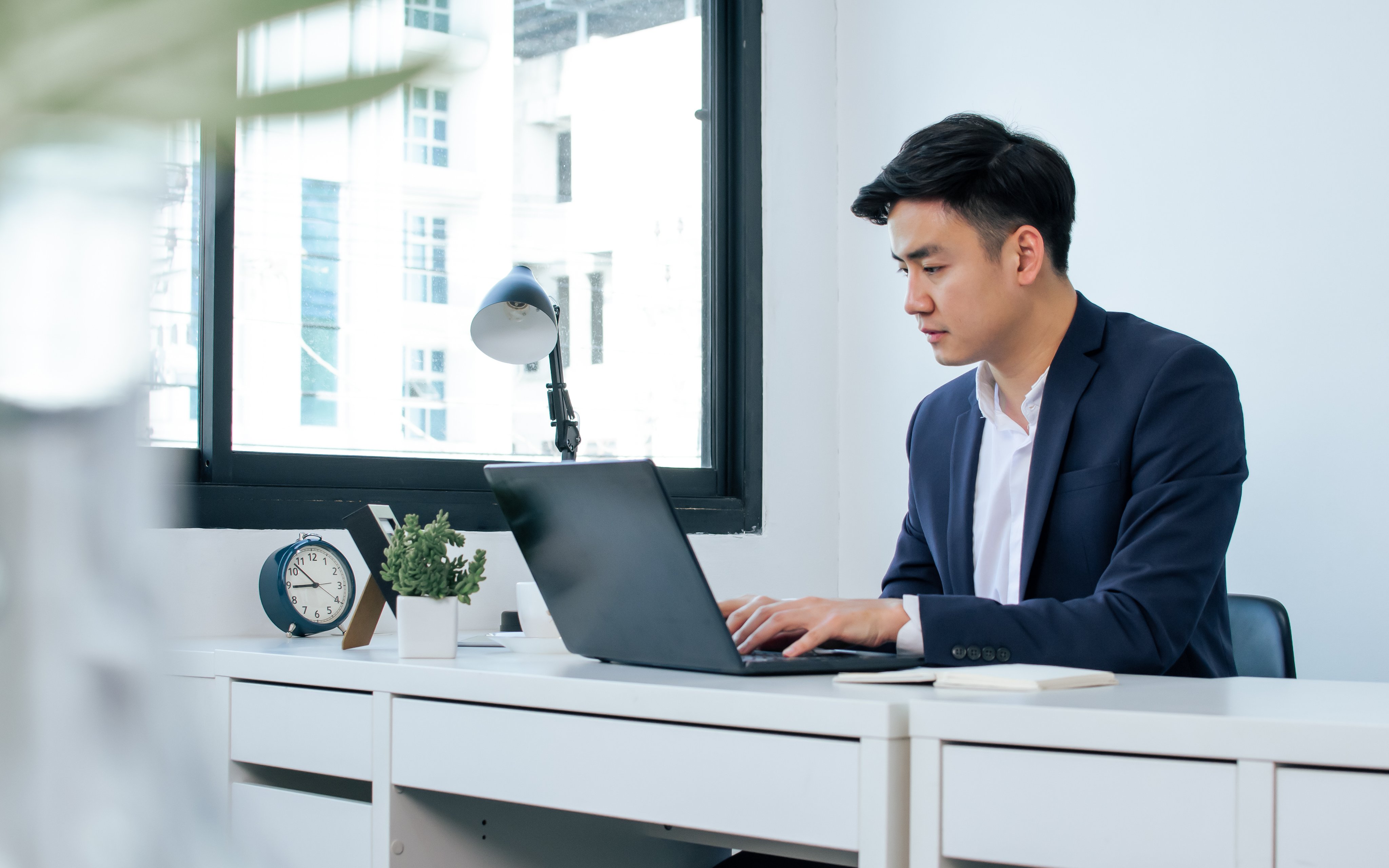 Asian handsome business man wearing white shirt and blue formal suit, communicating, working, typing on laptop, sitting near window in indoor office and smiling with happiness