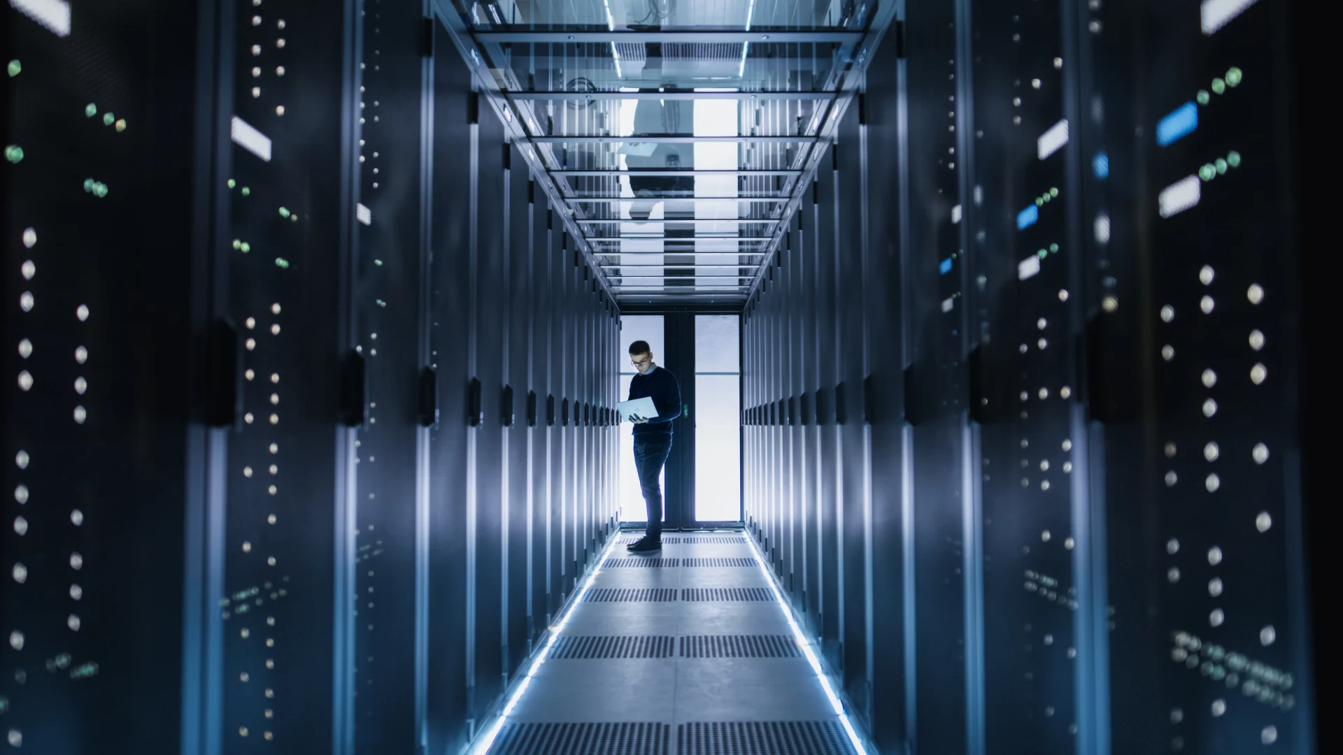 Male IT Engineer Works on a Laptop in a Big Data Center. Rows of Rack Servers are Seen.