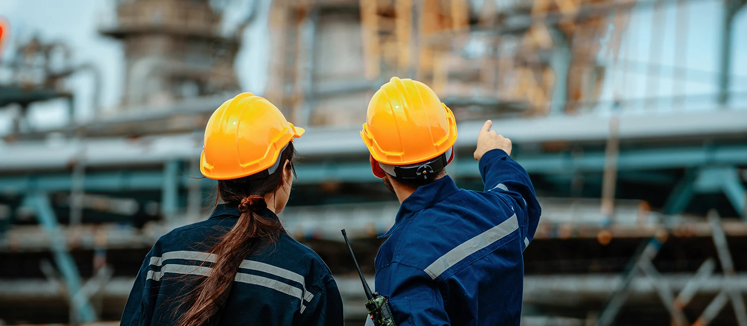 Two workers wearing hard hats examine machinery at an industrial facility. One points toward equipment while discussing safety protocols, emphasizing teamwork and attention to detail.
