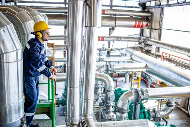 Refinery worker standing on metal bridge platform and controlling energy production inside heating plant.