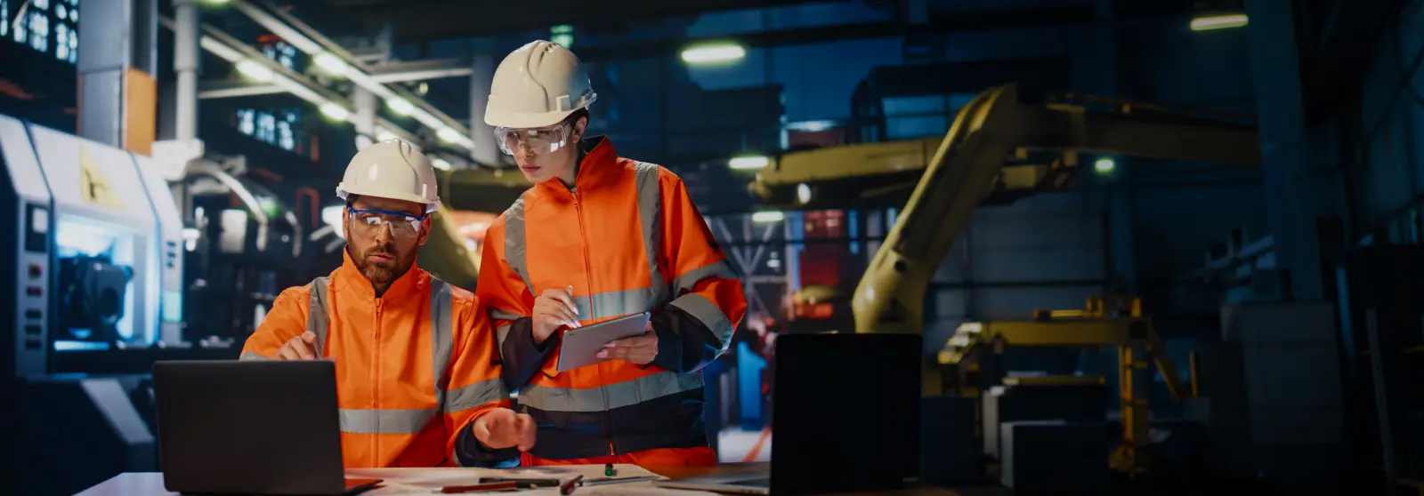Team factory workers talking together looking laptop computer at industrial manufactory. Two engineers in helmets uniform discuss innovation production. Couple professional specialists work on plant.
