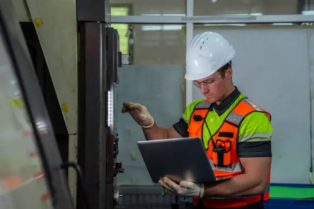 A man in a safety vest is looking at a laptop. He is wearing a hard hat. The laptop is on a table