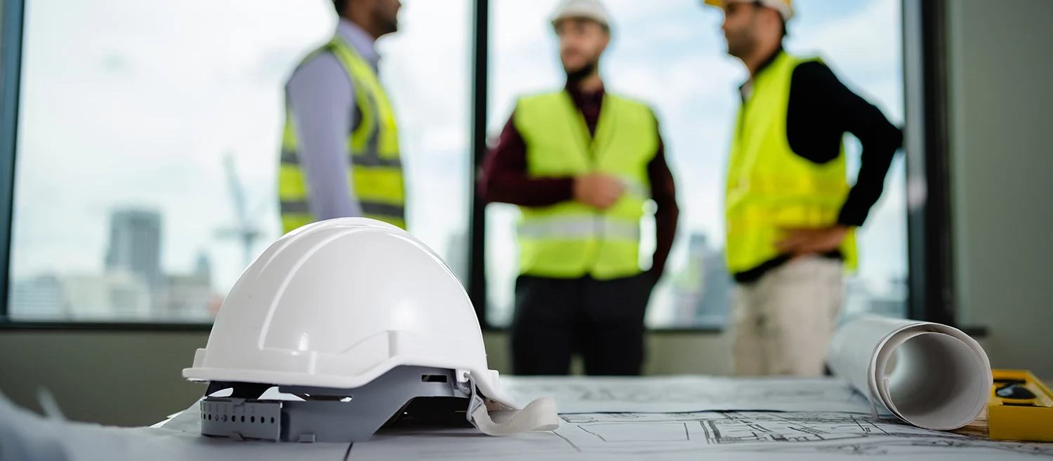 Selective focus of white helmet hardhat with a blueprint on the desk have a team of engineers diverse discussing in the background, architectural project, developer real estate, construction concept.