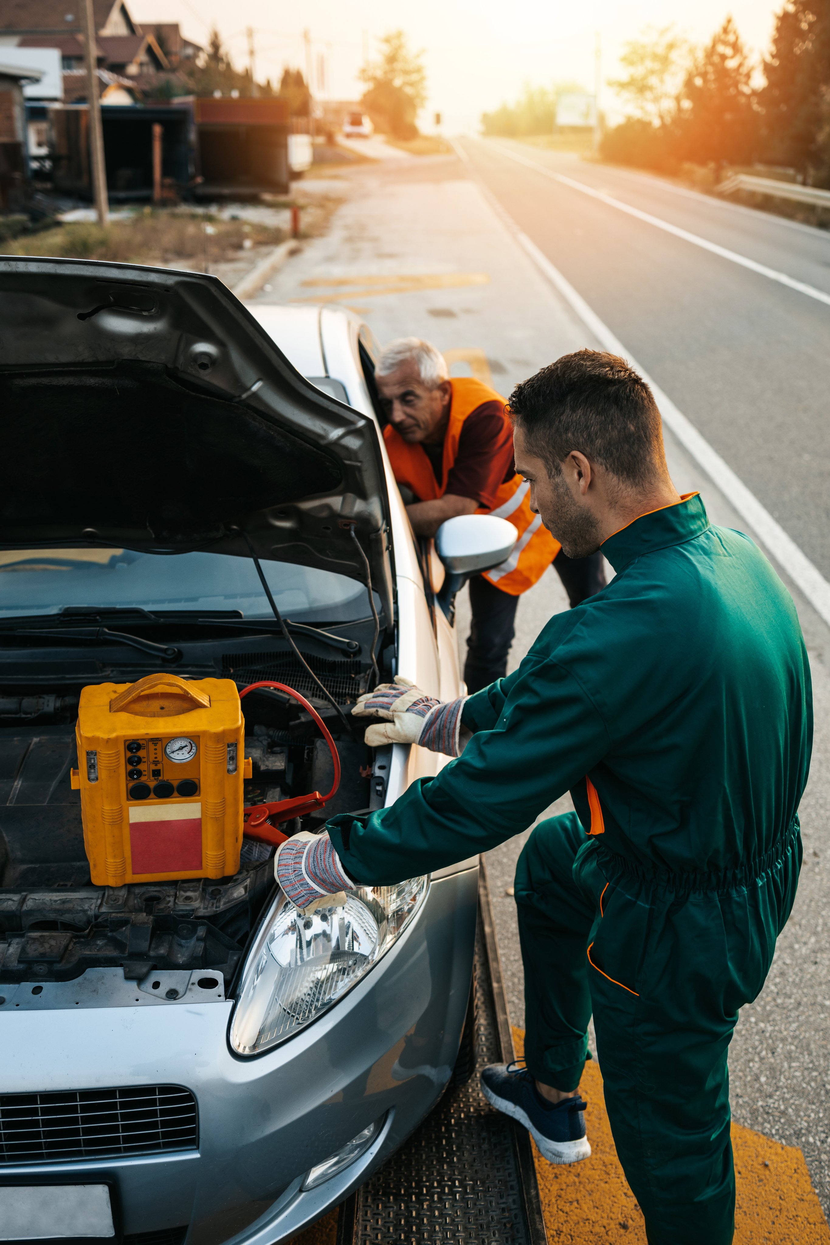 Two road assistant workers in towing service trying to start car engine with jump starter and energy station with air compressor. Roadside assistance concept.