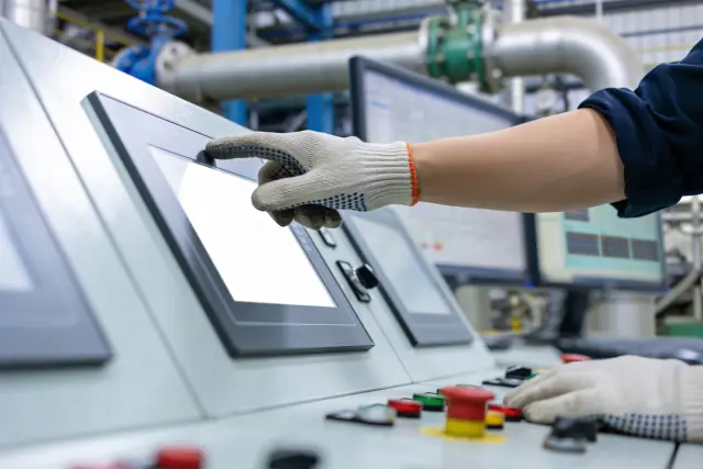 A worker wearing protective gloves touches a blank screen on a sophisticated control system in a factory setting, with monitors displaying data in the background