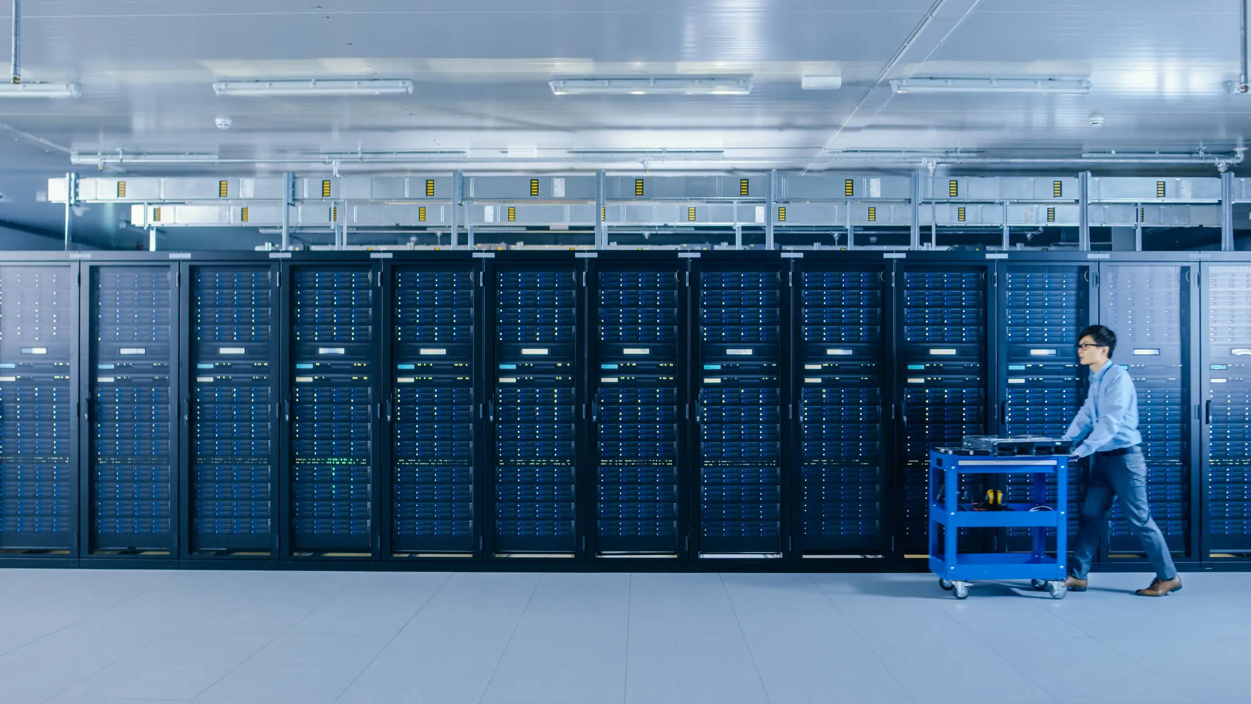 In the Modern Data Center: IT Technician Working with Server Racks, Pushes Cart Between Rows of Server Racks. On a Pushcart New Hardware for System Update. Engineer Doing Maintenance and Diagnostics.