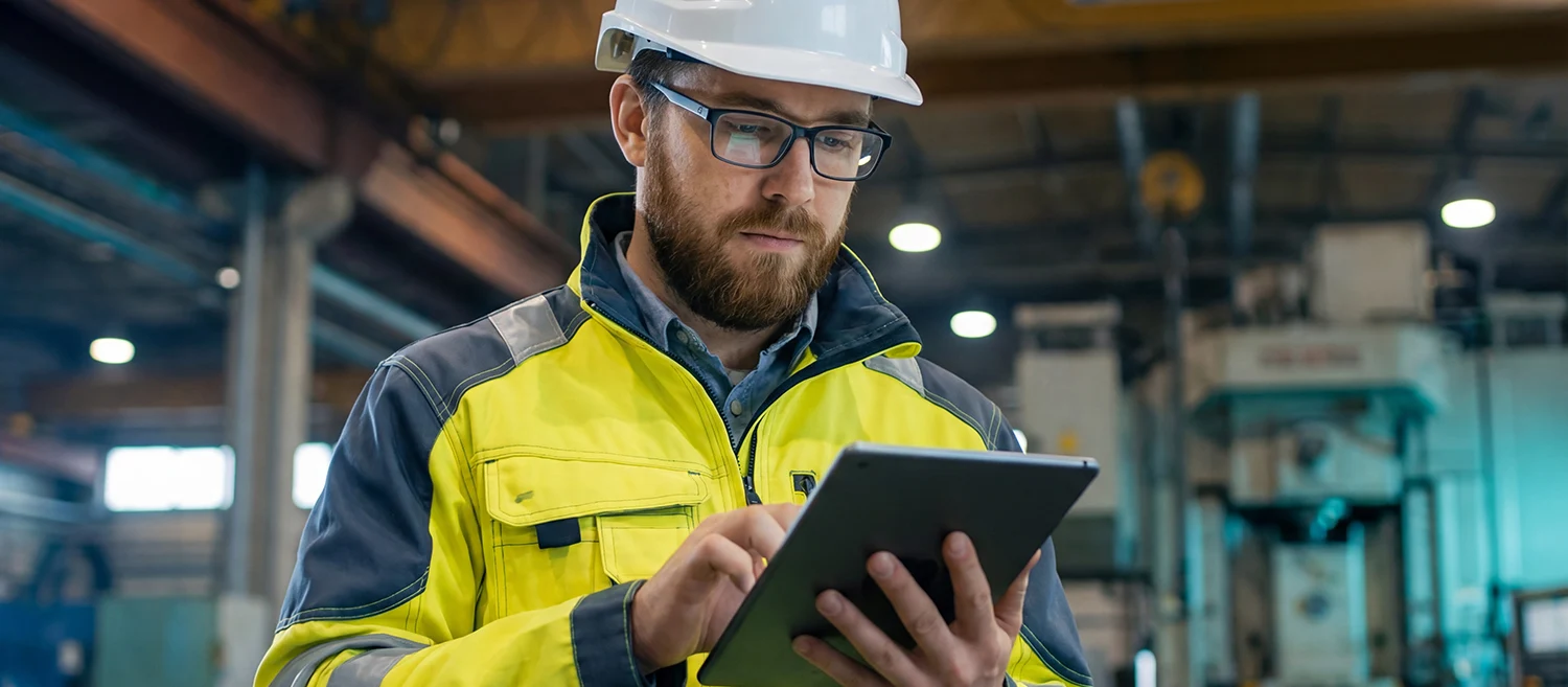 A bearded professional engineer in a high visibility jacket analyzes production data on a tablet device while standing on a busy factory floor