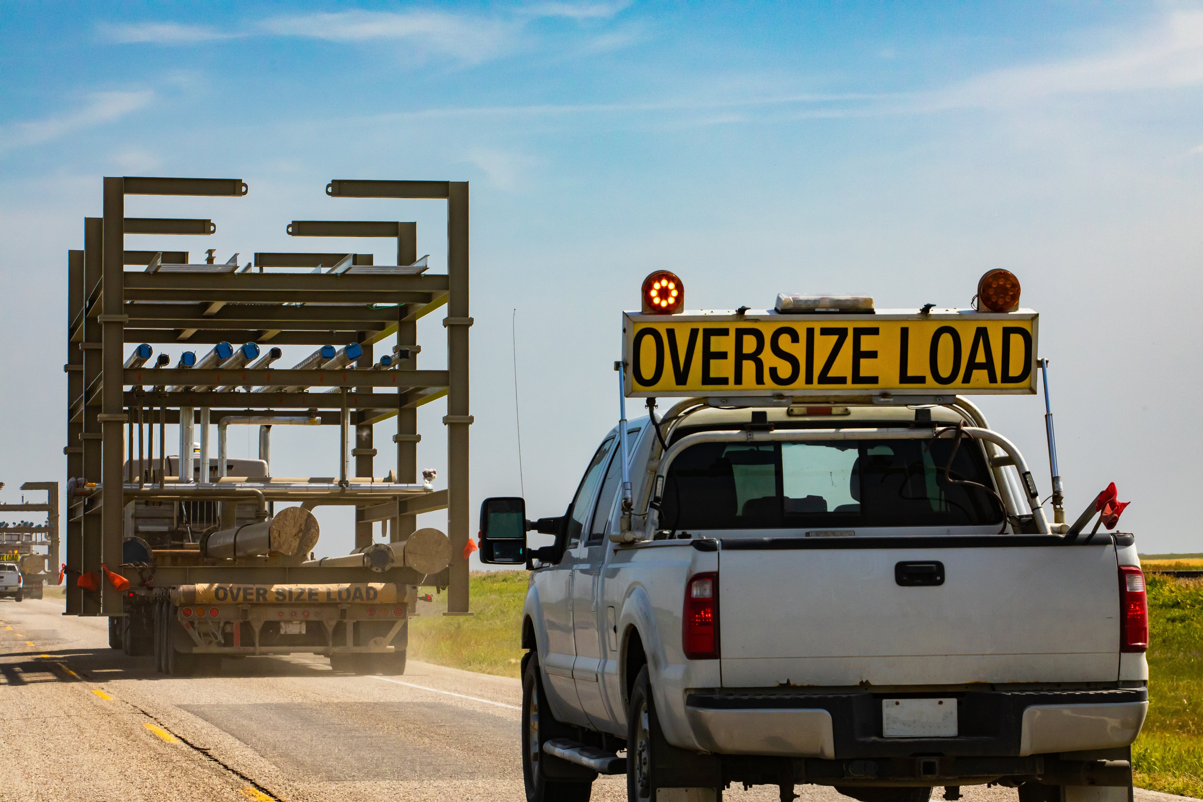 Oversized truck traveling along the highway on a sunny day. Pickup is following behind for security. Coutryside grasslands and plains in the background.