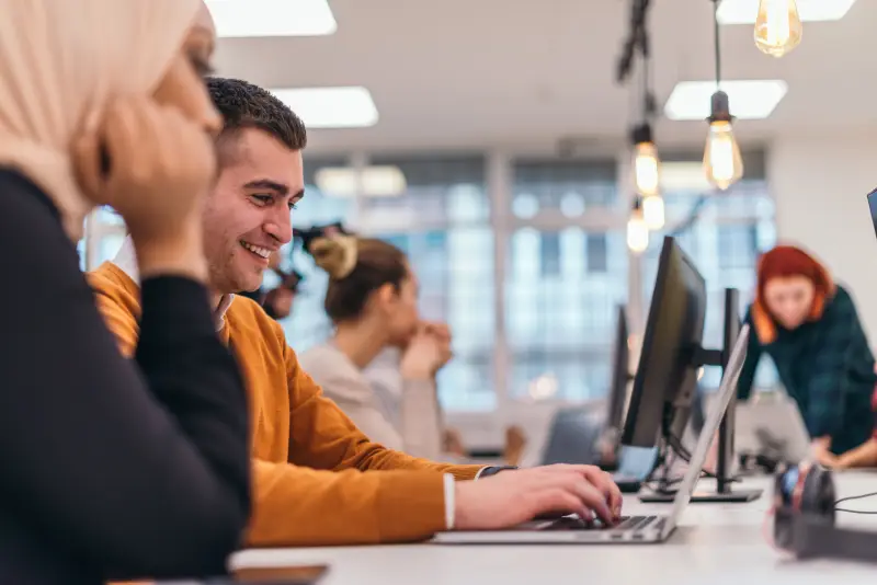 Close-up portrait of a happy young man sitting in an office next to his colleagues and working on his laptop.