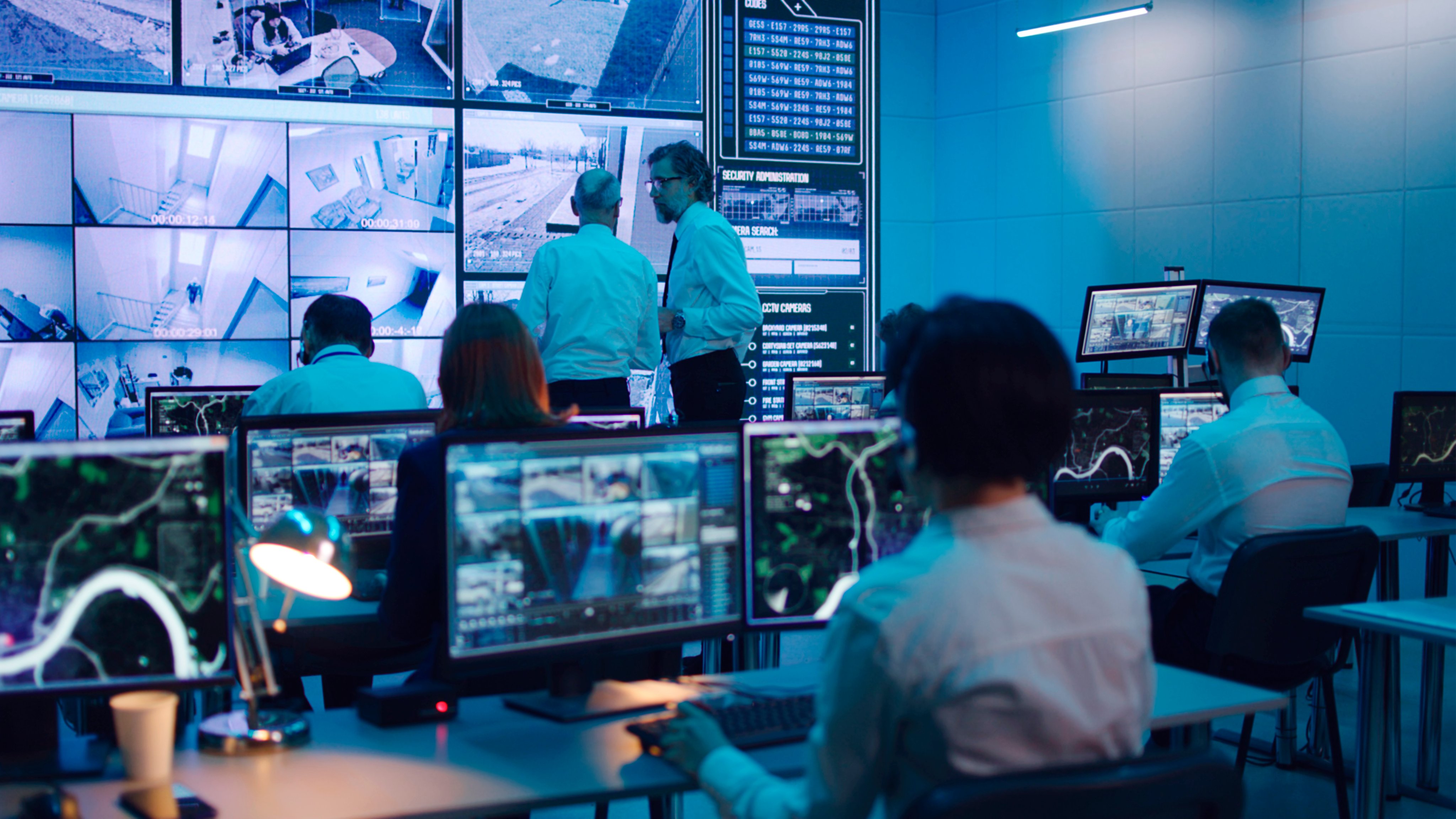 Mature men discussing surveillance footages on huge screen while male operator using computer during work in security center