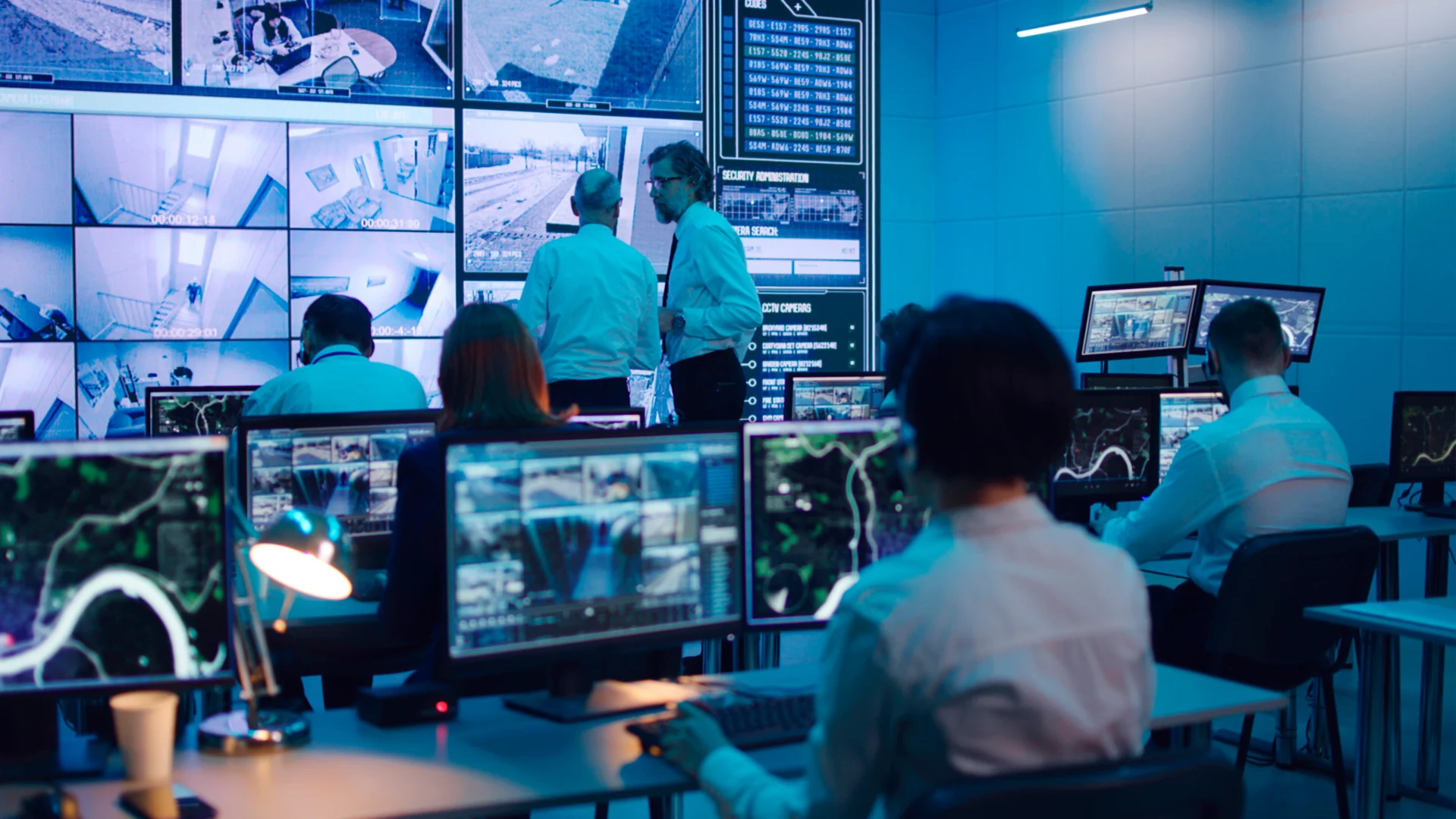 Mature men discussing surveillance footages on huge screen while male operator using computer during work in security center