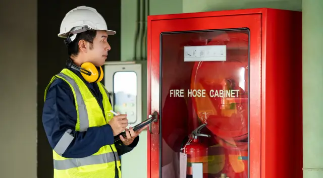 Building inspector man using digital tablet checking fire hose cabinet. Asian male worker in green vest, ear muffs and safety helmet working for building maintenance inspection