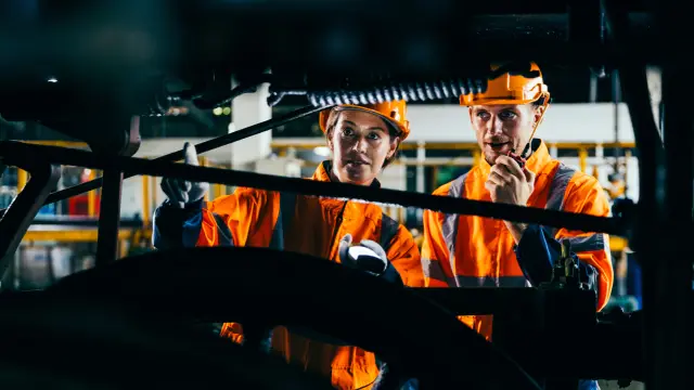 Two engineers wearing safety gear and helmets inspect machinery under train in railroad maintenance facility, showcasing teamwork, mechanical engineering and railway system inspection.