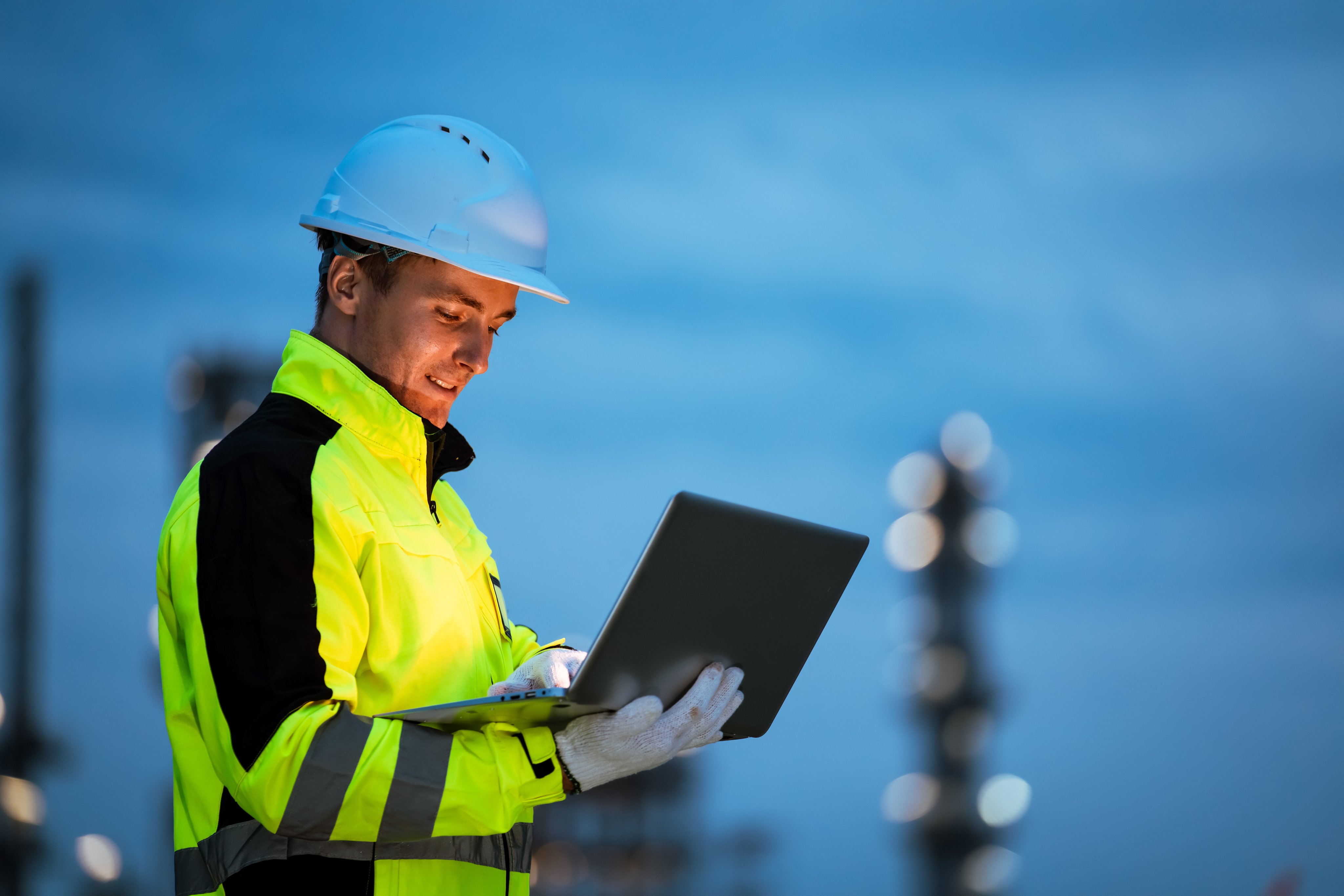 Factory worker engineer man using laptop computer for system maintenance at outdoor refinery site during night shift, working in industrial environment with focus and precision.