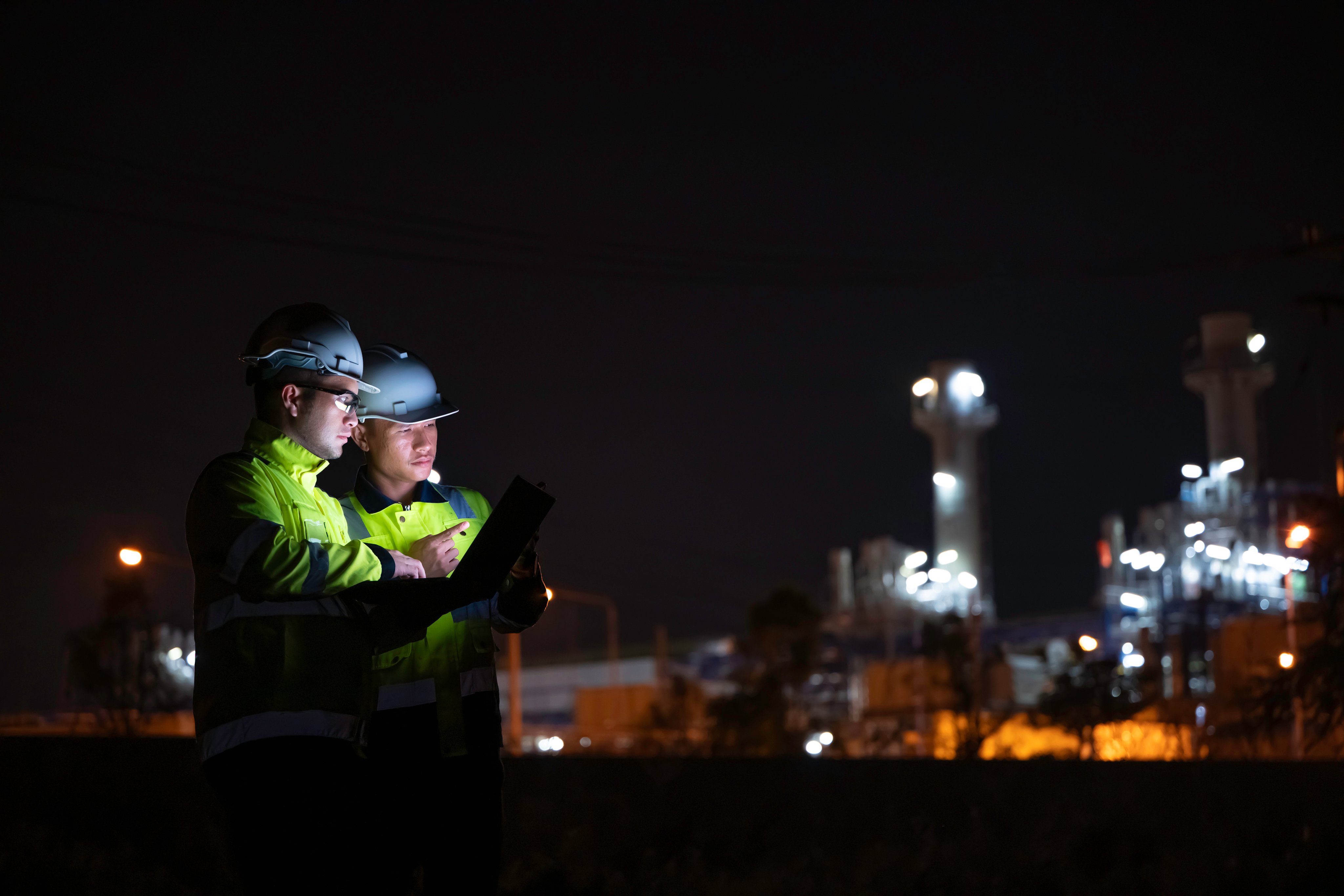 Industrial engineer night inspection at refinery or petrochemical plant walkie-talkie hand and digital tablet intersection of technology, worker work at night at power plant