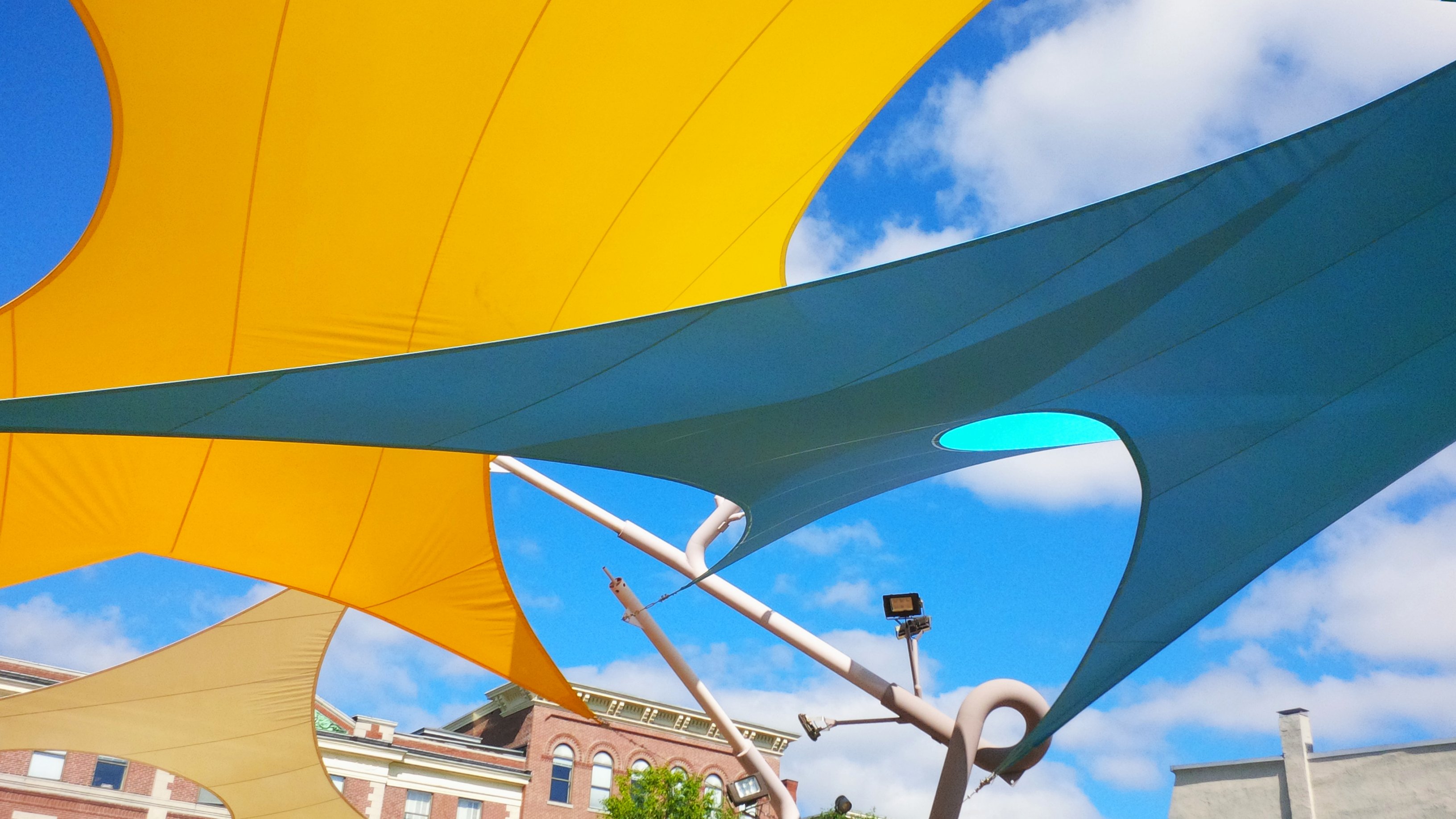 Overlapping yellow, teal and tan shade sails on angled steel masts against a blue sky