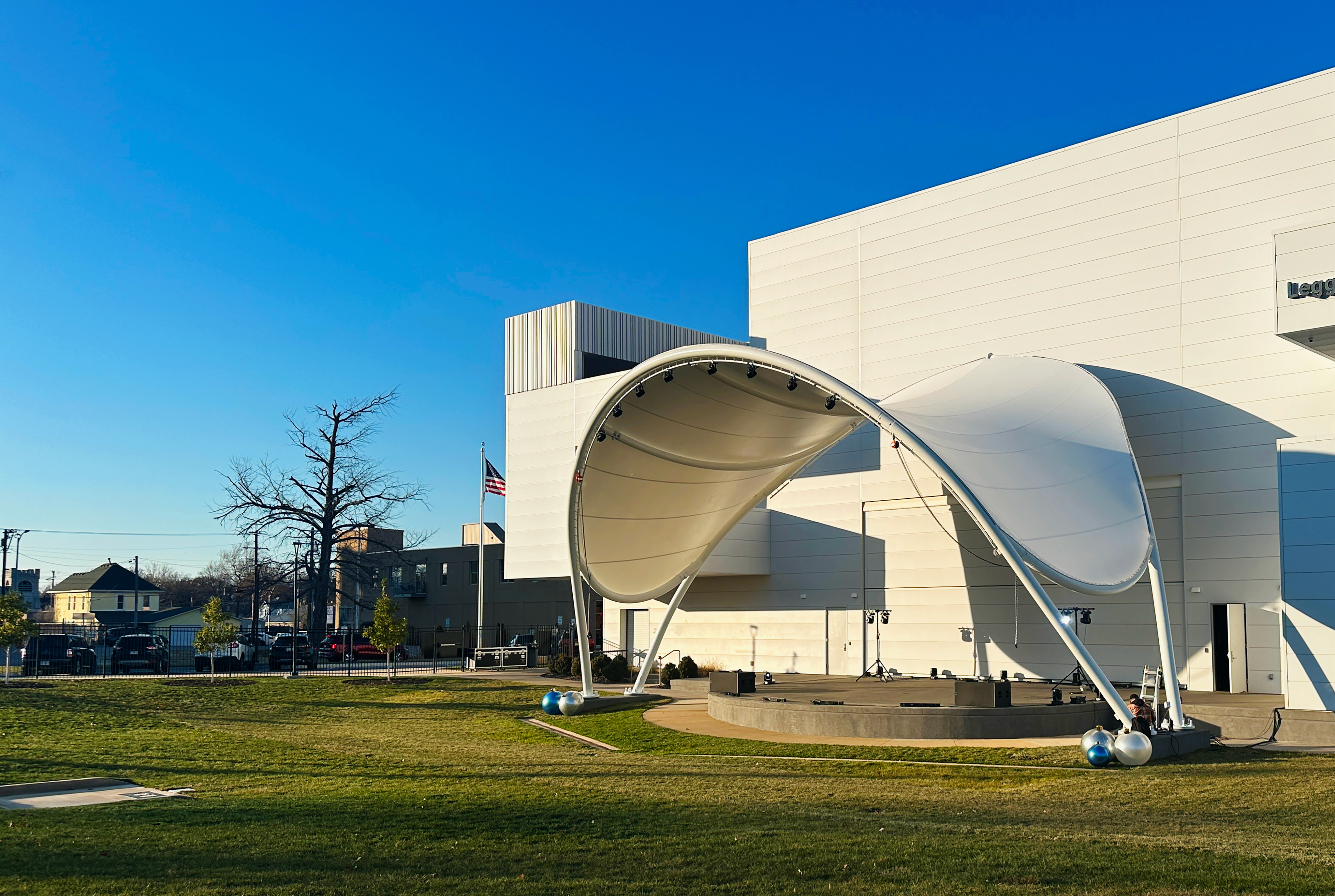 Tensile fabric stage canopy on an arched steel frame beside a white building