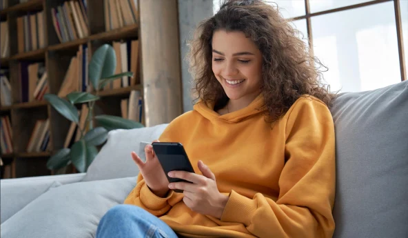 Person in a hoodie smiling at phone on sofa.