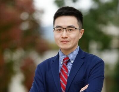 Michael Chi stands with arms crossed in front of greenery, wearing a suit and glasses
