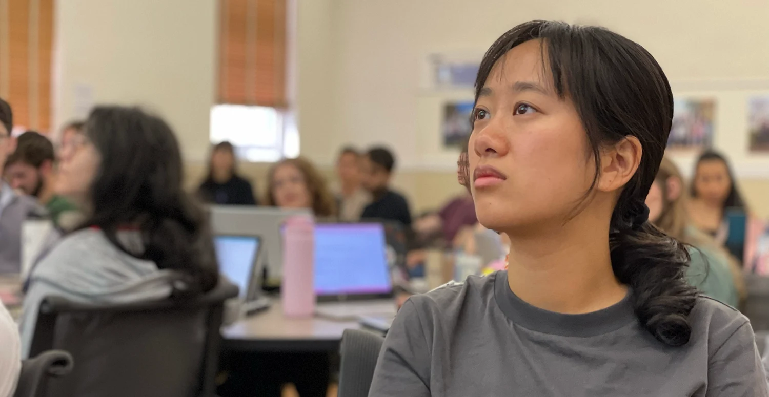 Student looking ahead at projection screen (not pictured) in classroom; other students are blurred in the background.