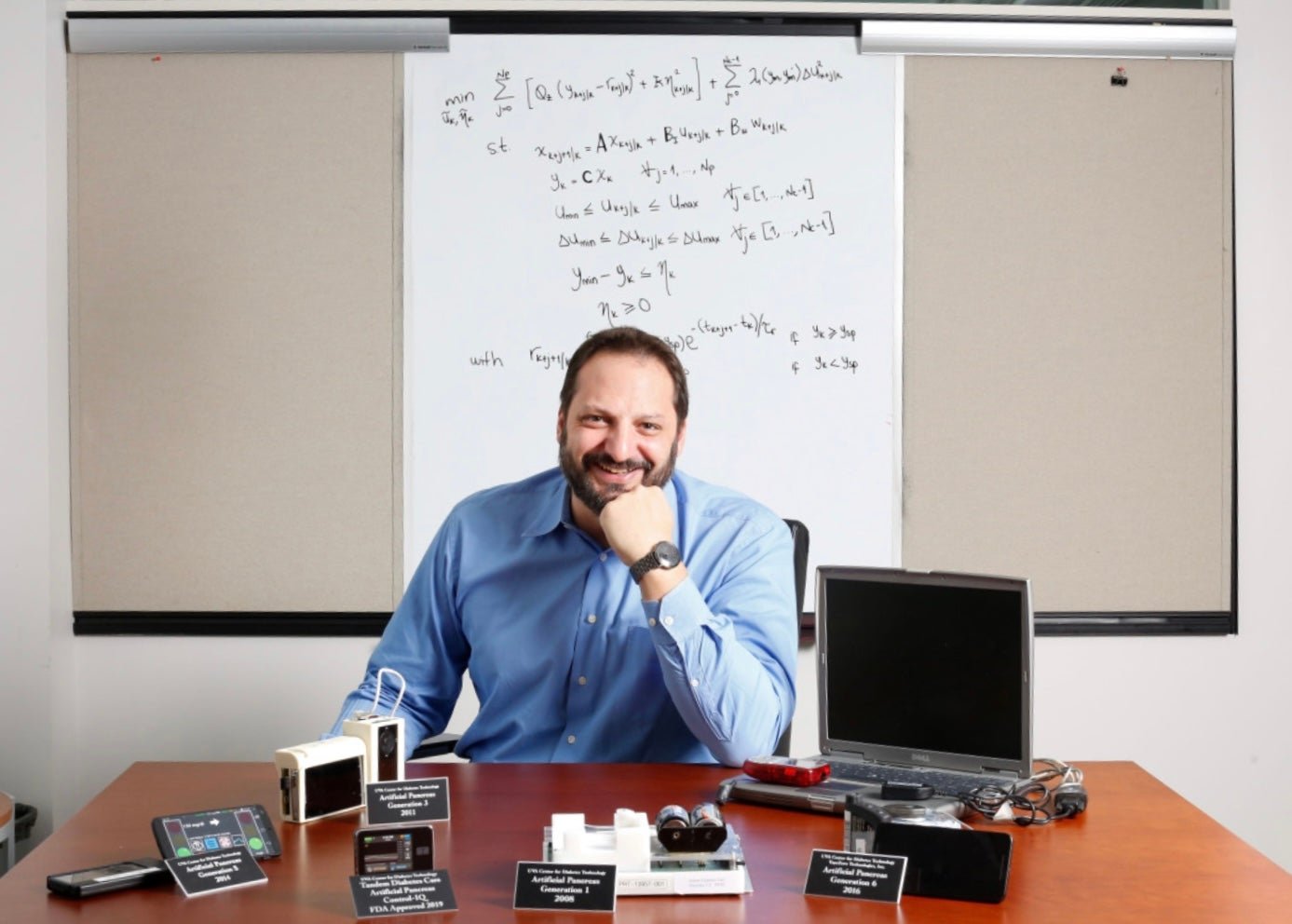 University of Virginia Center for Diabetes, Marc Breton PhD, photographed in an office setting with all generations of the artificial pancreas on the desk in front of him, also with an equation on a white board behind him.